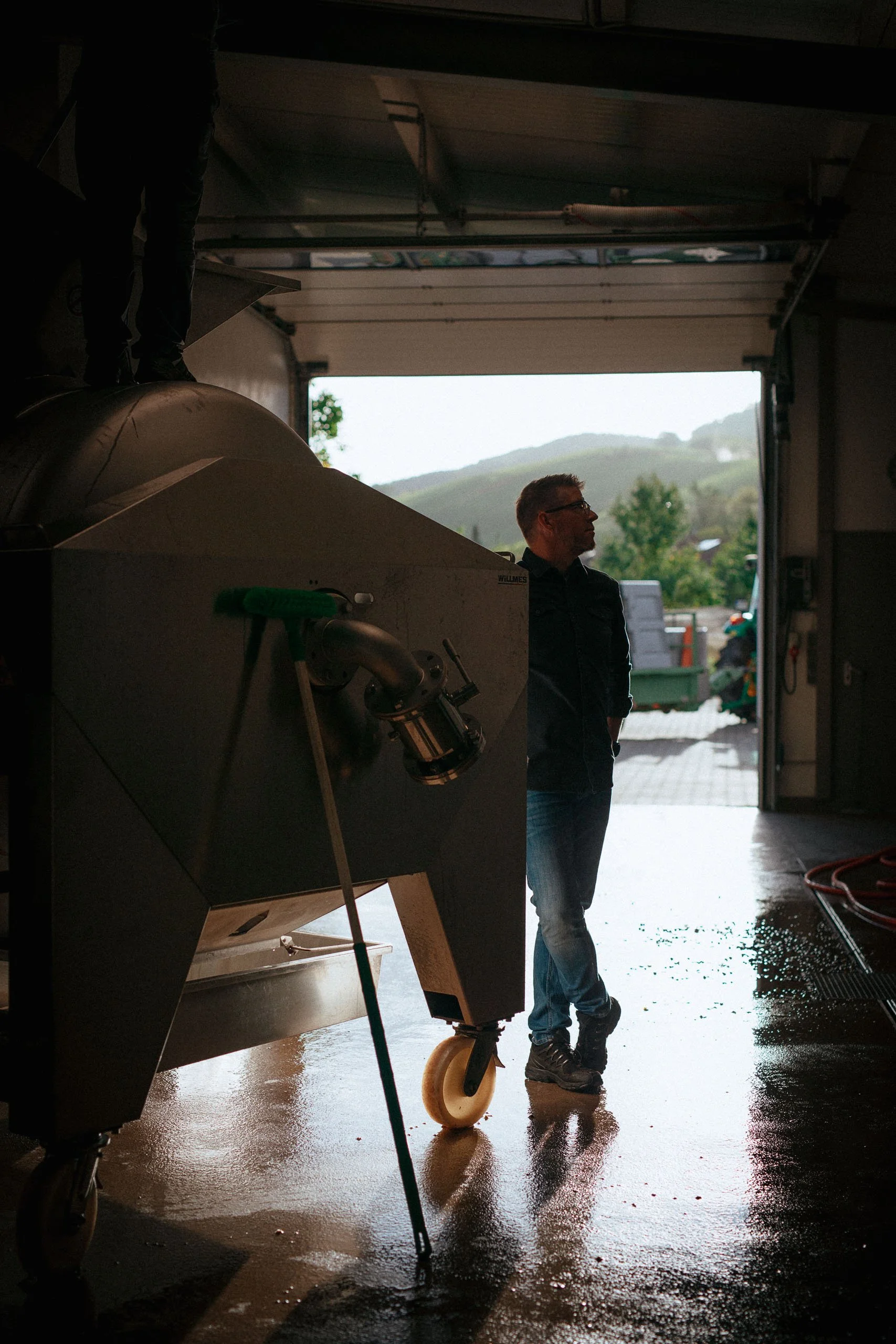 Mann steht in einer Garage neben landwirtschaftlicher Maschinen, im Hintergrund ein offener Ausgang zeigt eine ländliche Landschaft mit Hügeln und Bäumen, der Boden ist nass.