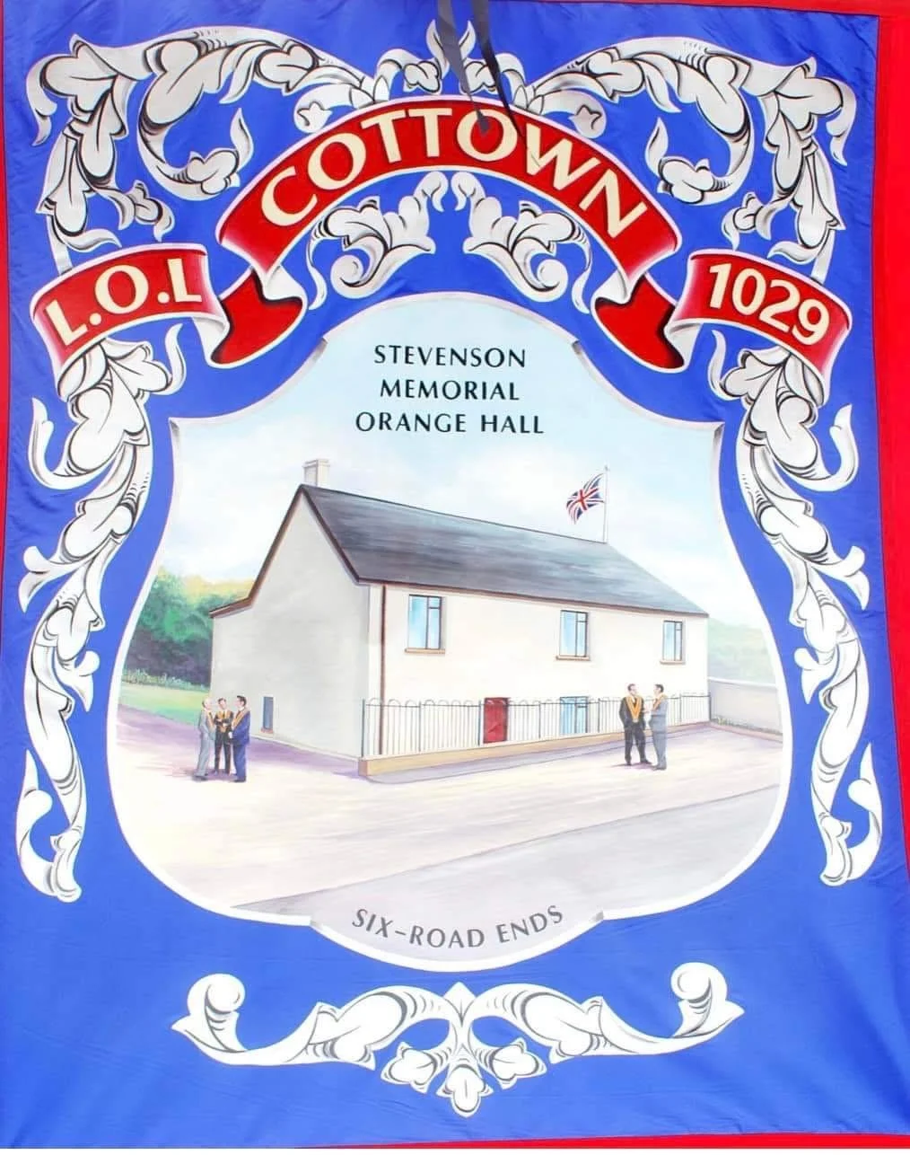 A decorative sign featuring a painted illustration of the Stevenson Memorial Orange Hall, a white building with a gray roof and a Union Jack flag, surrounded by ornate blue and white scrollwork, and text indicating the location as Cottonown, with the postcode L.O.L 1029, and the phrase 'Six-Road Ends.'