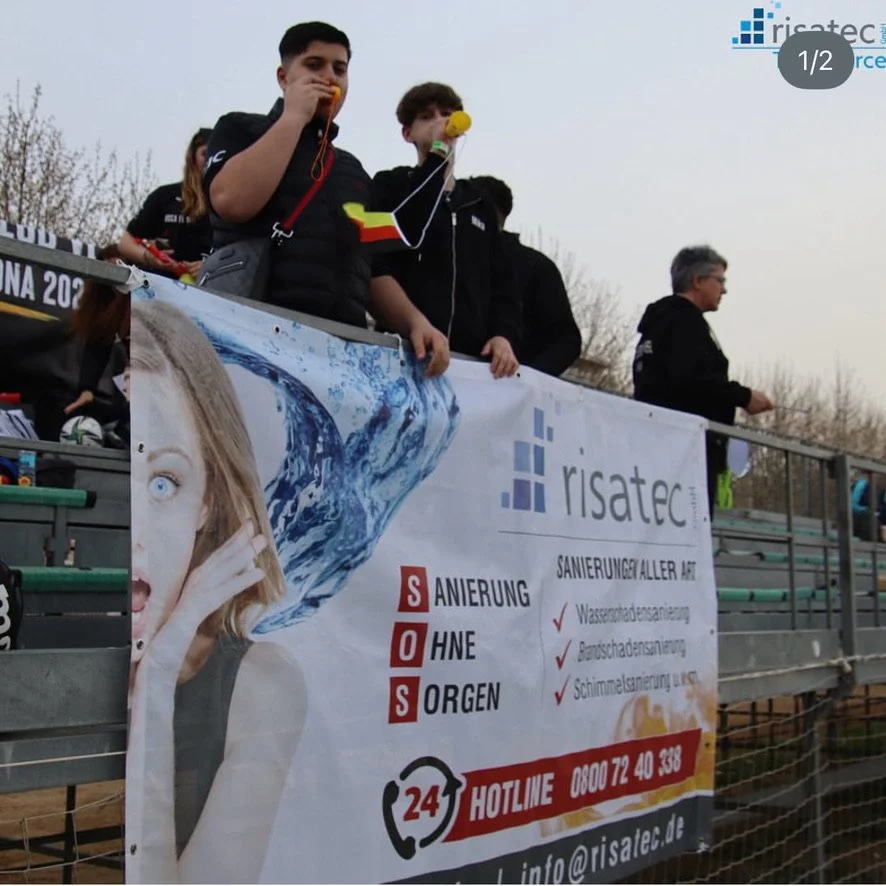 Sponsoring: Menschen stehen auf einer Tribüne, einige blasen in Tröten, dahinter ein Banner mit Werbung für risatec GmbH. Wasserschadensanierung, Bauschadensanierung und Schimmelpilzsanierung und vieles mehr.
