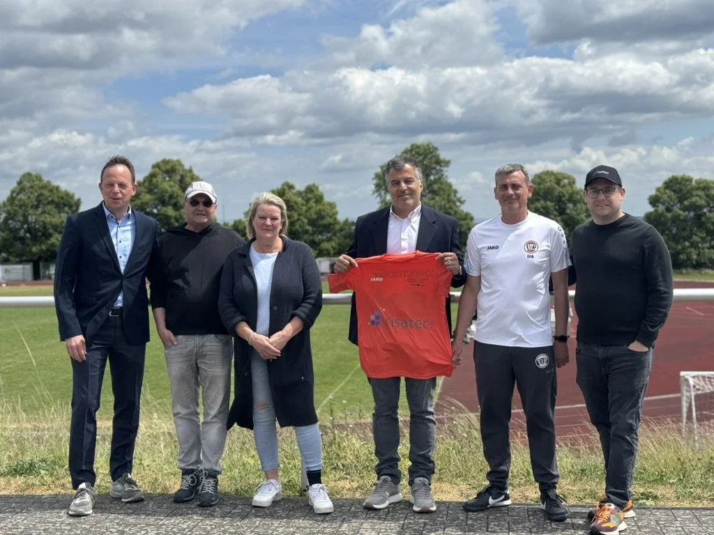 Sechs Personen stehen nebeneinander auf einer Laufbahn im Freien, einige halten eine rote Sportschüte. Der Himmel ist bewölkt.