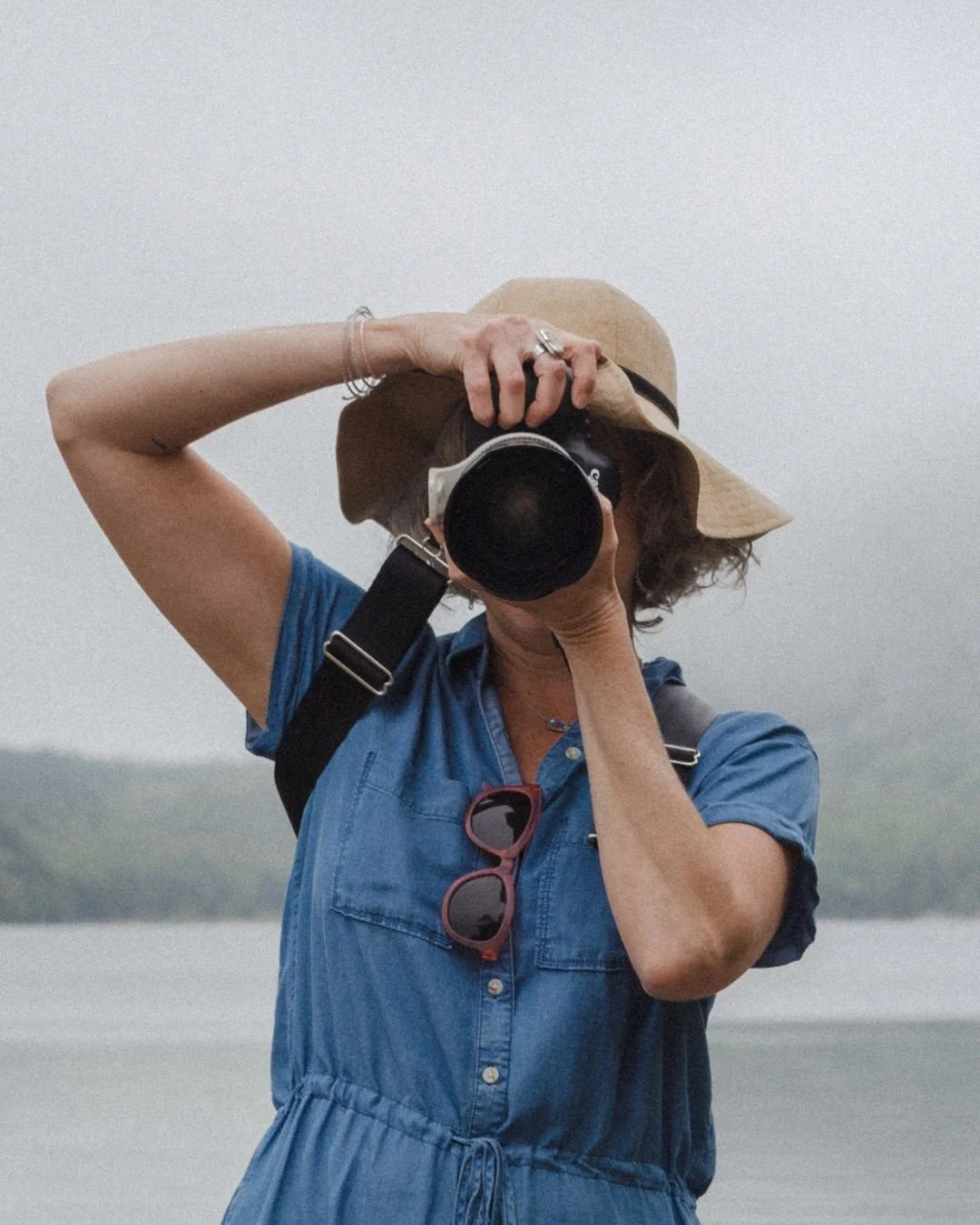 Person taking a photo with a camera near a body of water, wearing a wide-brimmed hat, sunglasses hanging from the shirt, and a blue dress.