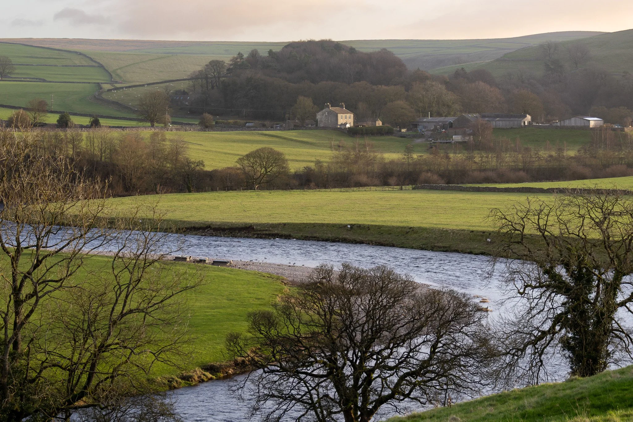 Scenic rural landscape with a winding river, green fields, leafless trees, and farm buildings in the distance under a cloudy sky.