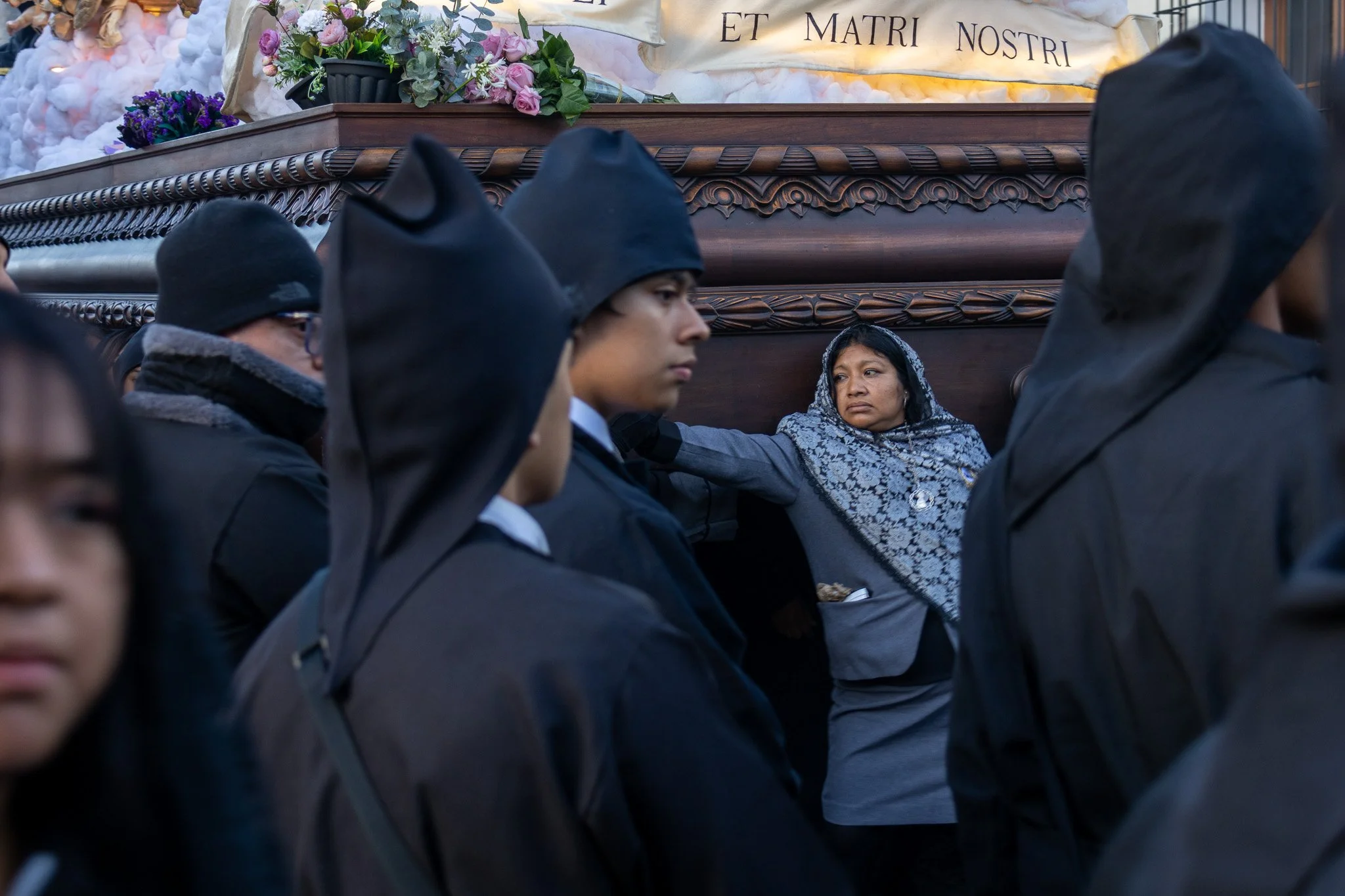 People wearing black clothing and head coverings are standing close together in a somber scene, with a woman in headscarf sitting behind a dark wooden structure adorned with flowers and a Latin inscription.
