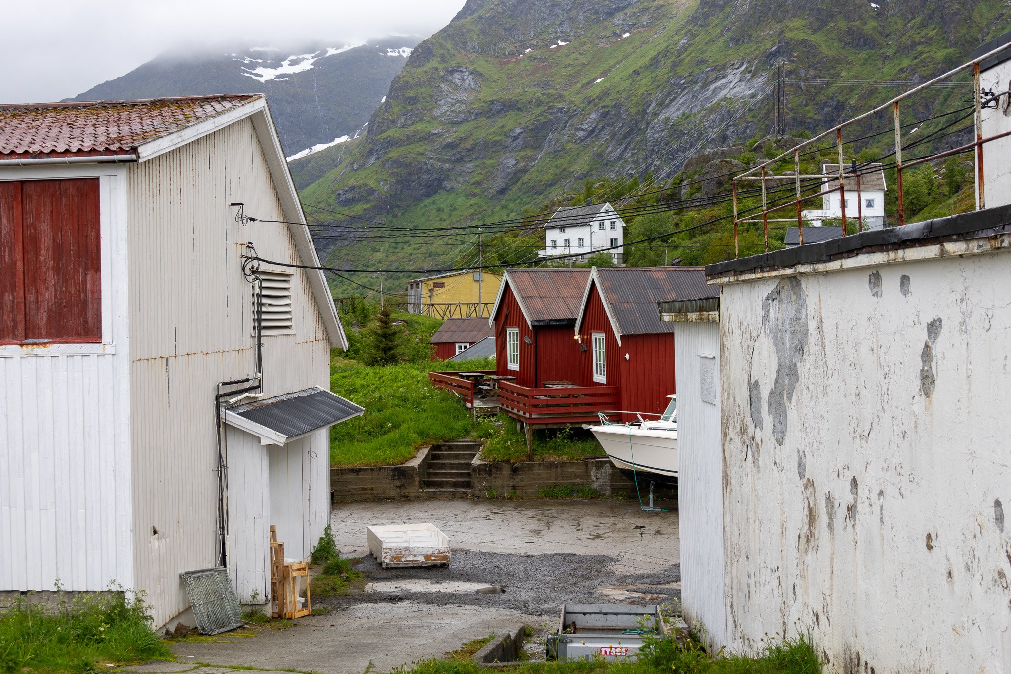 View of a rustic seaside village with colorful small houses, some on stilts, set against green mountains and overcast sky. There is a boat docked near white buildings, with outdoor stairs leading to a grassy area.