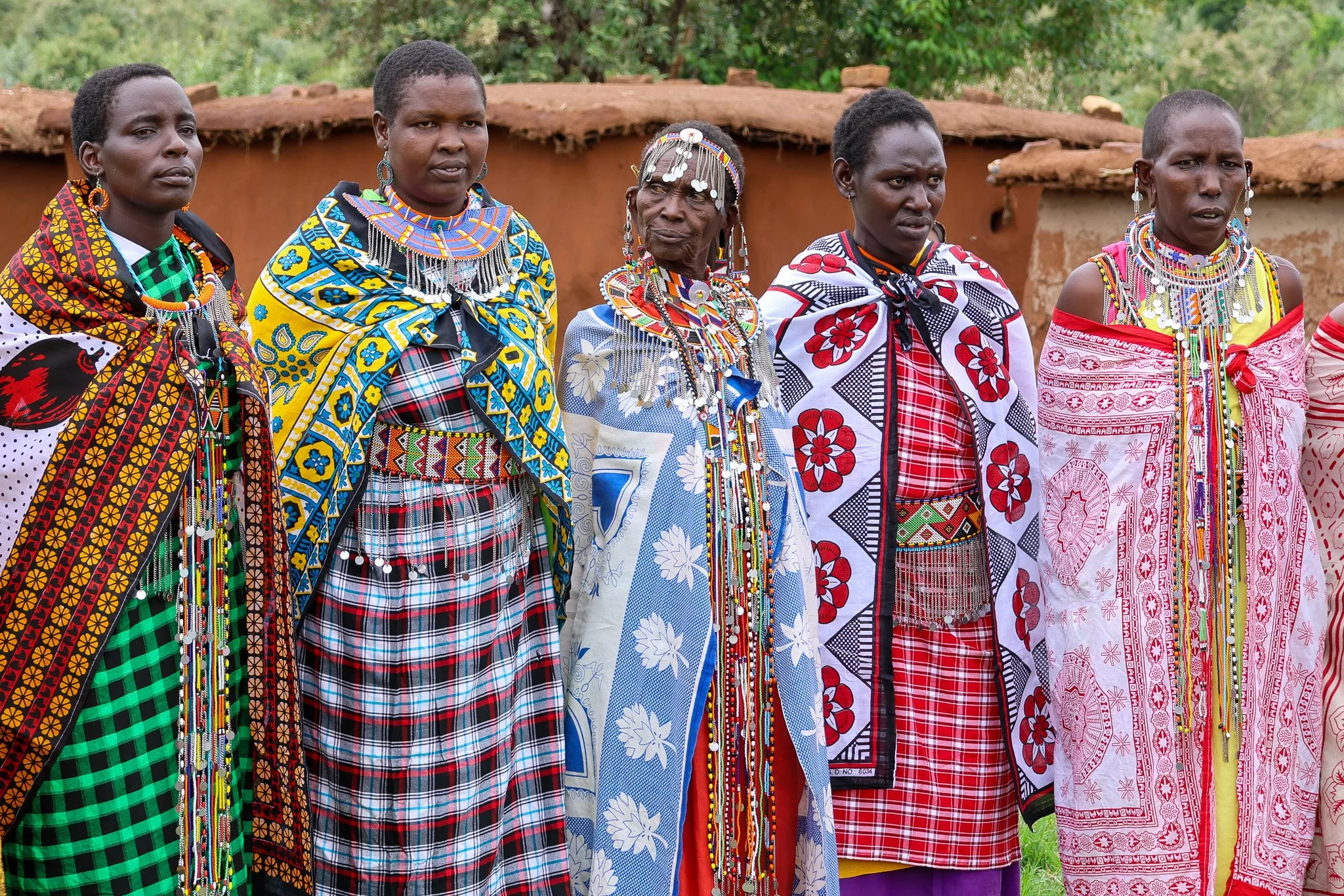Five women in traditional Maasai attire and jewelry standing outdoors in front of a mud structure with greenery.