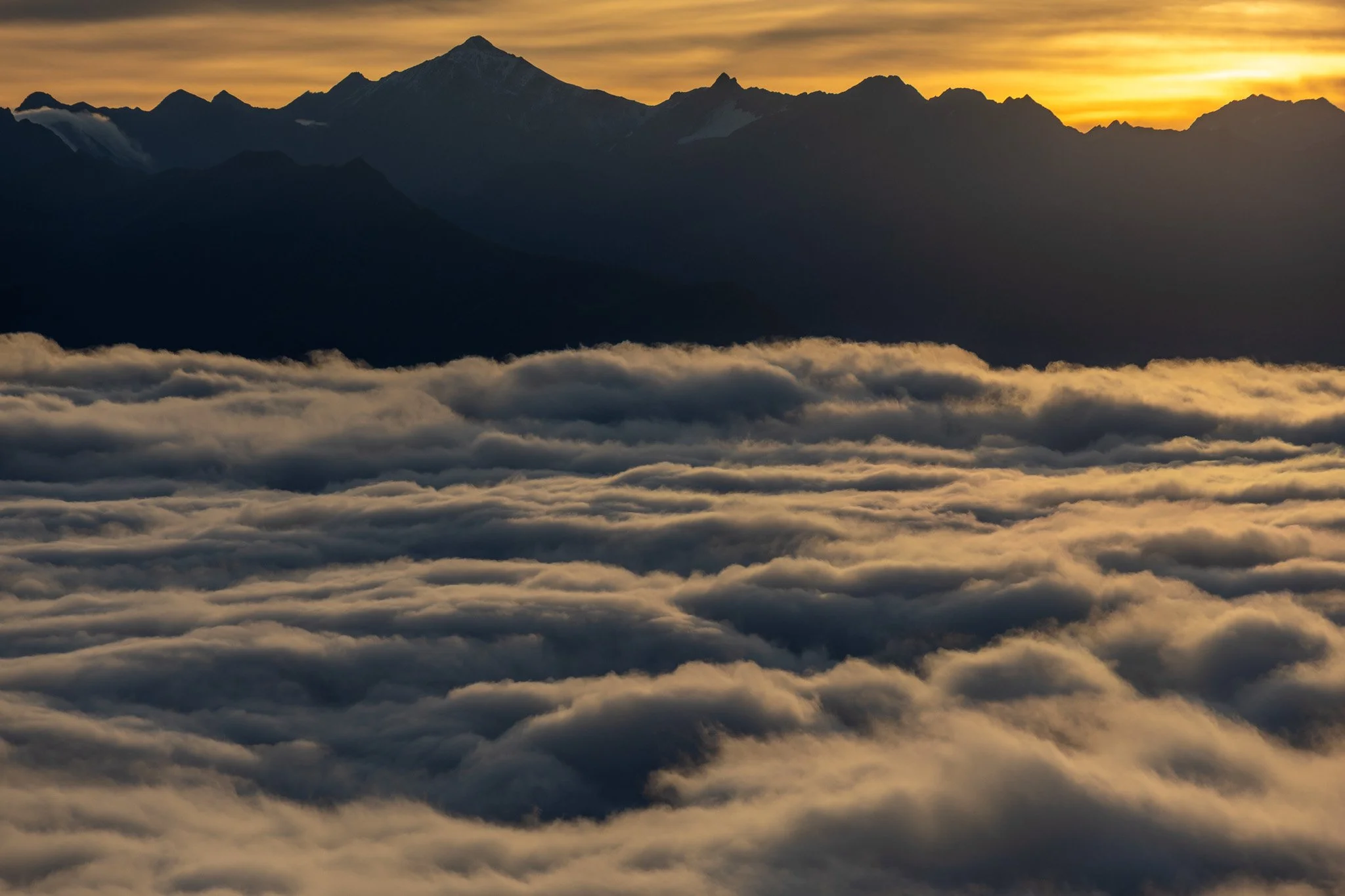 A mountain range at sunrise, with the peaks silhouetted against a golden sky, and a layer of clouds below covering the landscape.