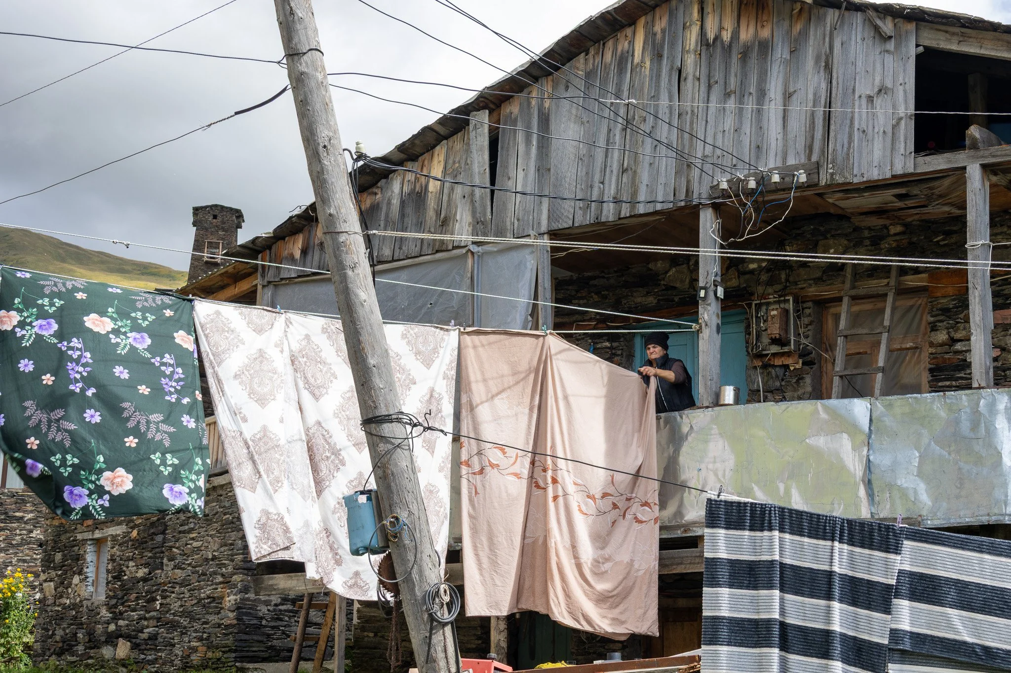 A woman hanging laundry on a clothesline outside a rustic, two-story wooden house with a stone foundation. Several pieces of fabric and clothing are hanging, and a ladder leans against the house. Overcast sky in the background.