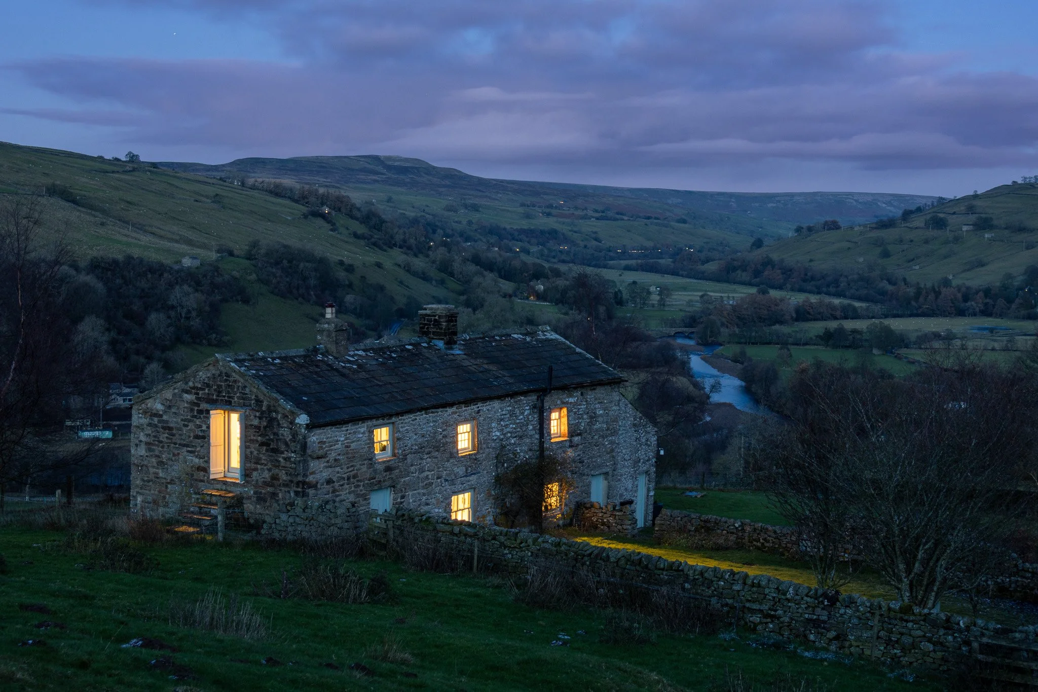 A stone house with lit windows at dusk, overlooking rolling green hills and a river in a rural landscape.