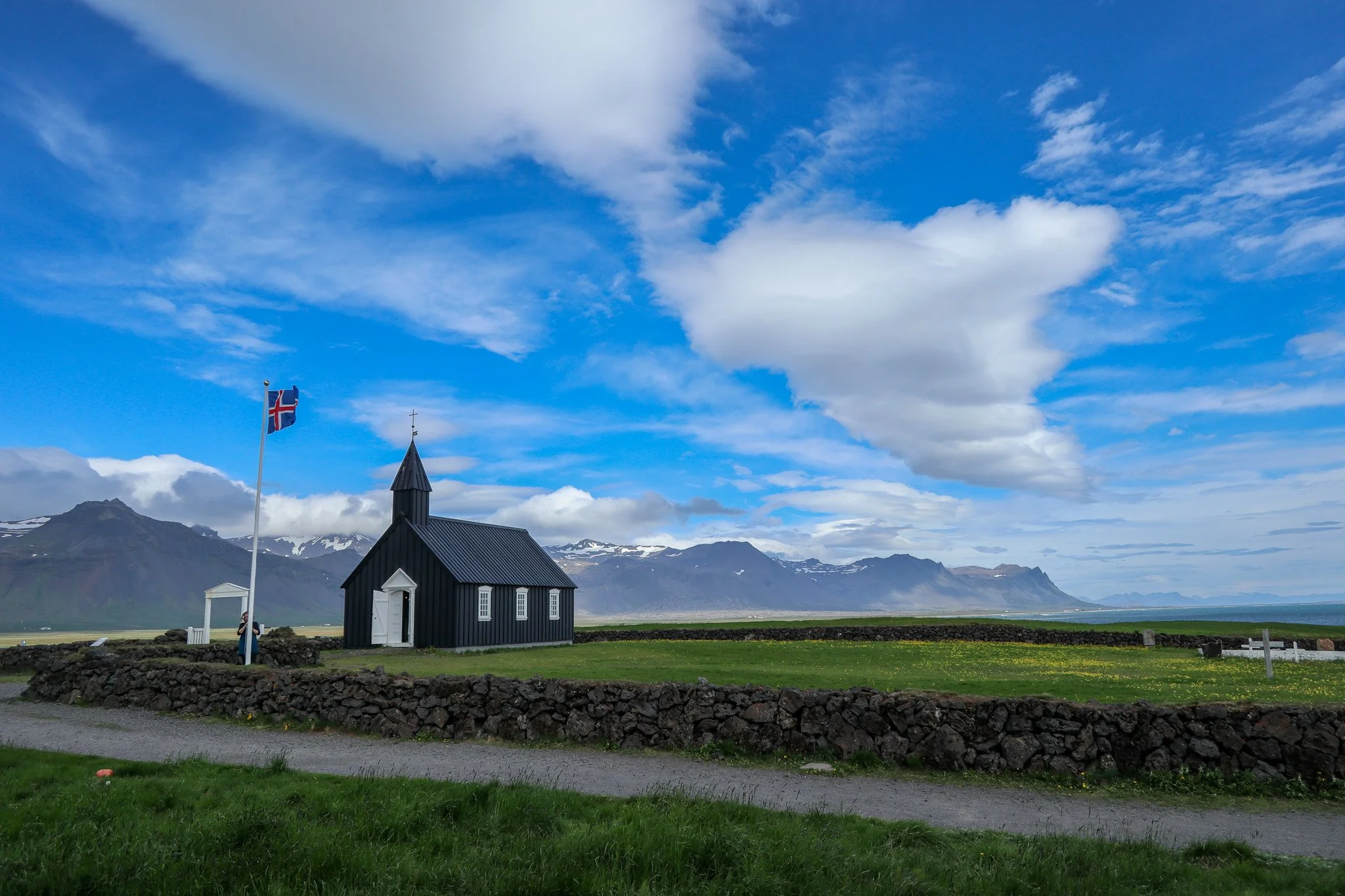 A small black church with a white door and windows, a flagpole with a Norwegian flag, stone wall, green grass, mountains with snow patches in the distance, blue sky with scattered clouds.