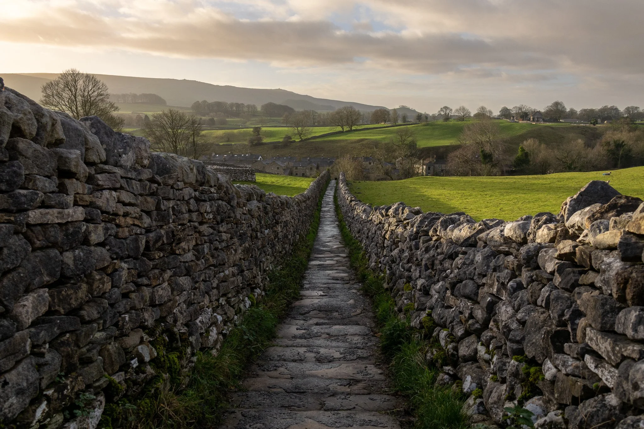 A narrow stone pathway flanked by dry stone walls, leading toward a lush green landscape with rolling hills and a small village under a cloudy sky.
