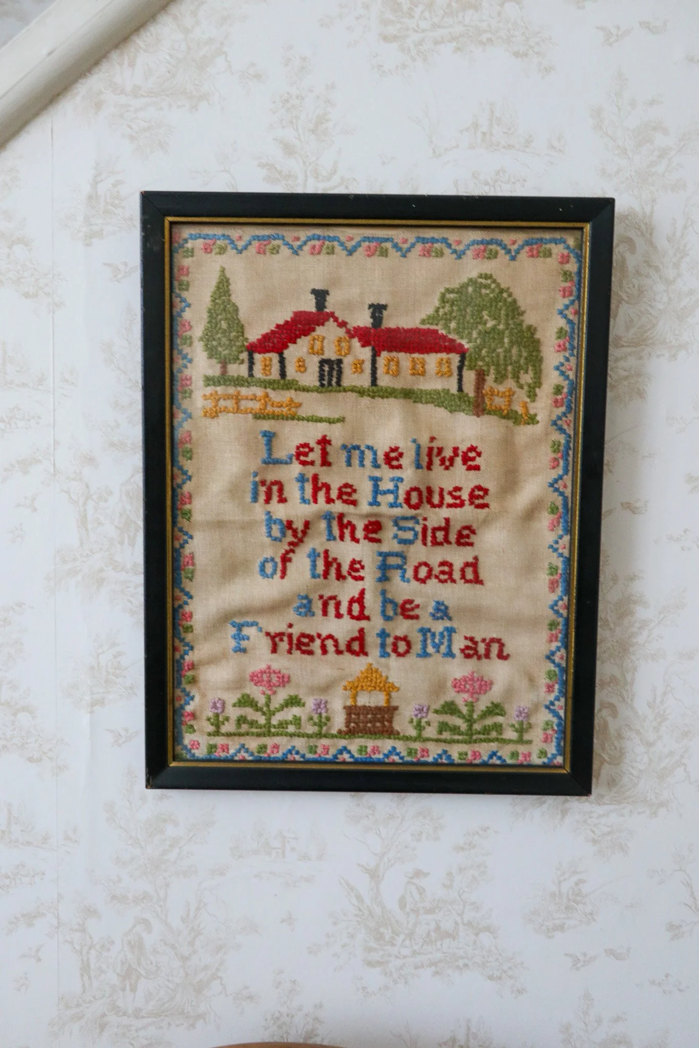 Frame with embroidery of a house, trees, flowers, and a quote reading "Let me live in the house by the side of the road and be a friend to man."