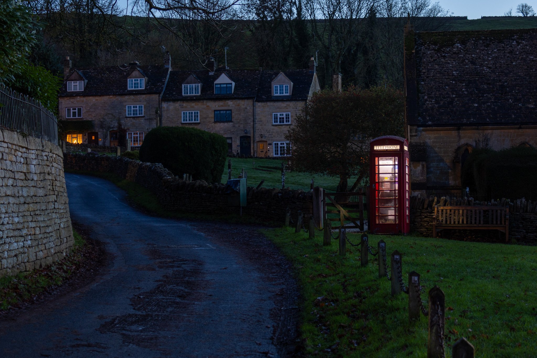 A narrow, winding road in a small village at dusk or night. On the right side, there is a classic red telephone booth and a wooden bench. The background shows old stone houses with illuminated windows, surrounded by trees and a hillside.