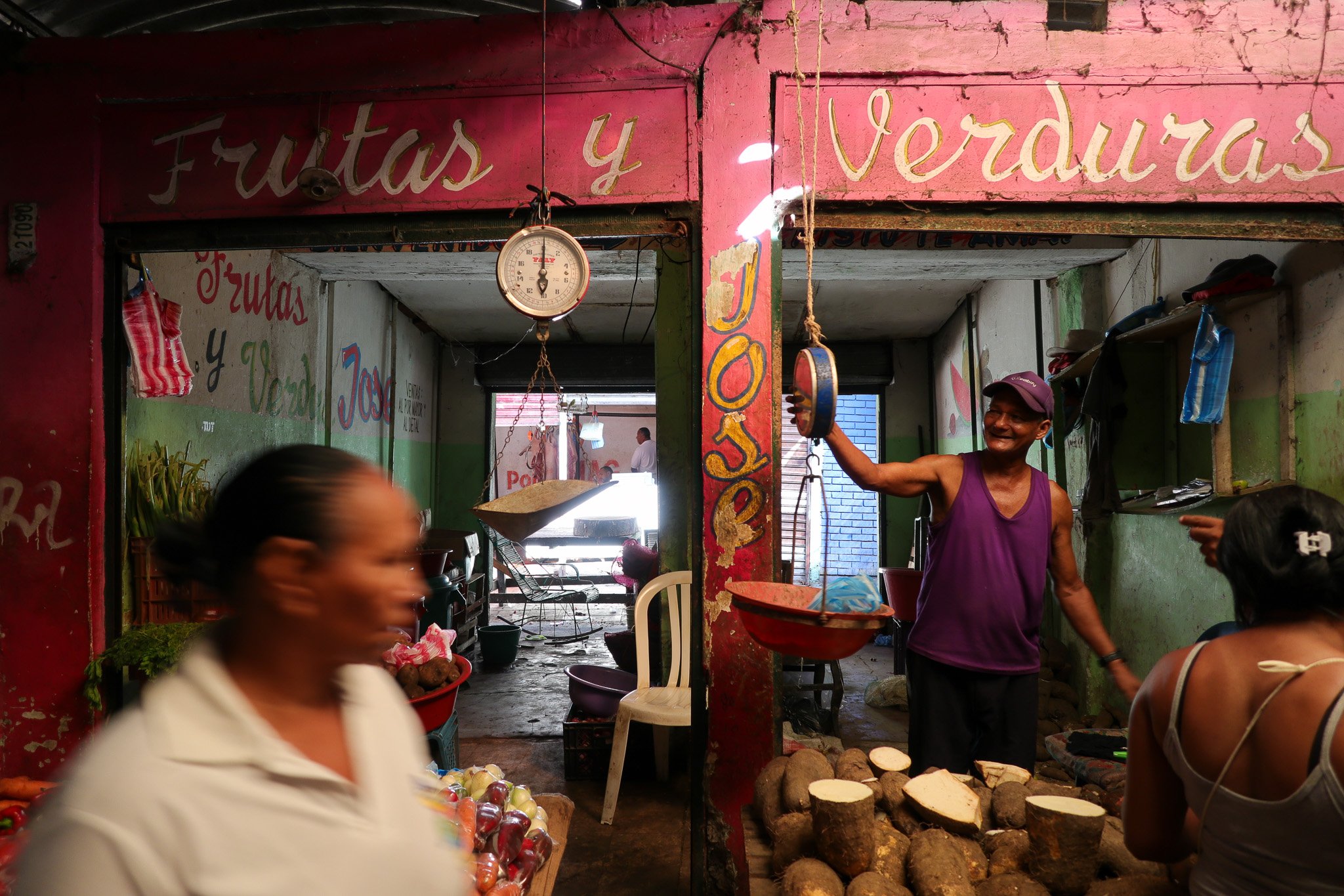 A man wearing a purple tank top and cap smiling and weighing yuca root at a small market stall with a woman in front of him, blurred face, and another woman with black hair and a white tank top pointing at him.