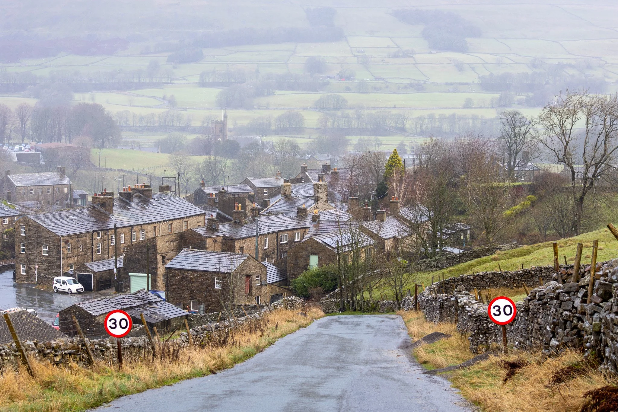 A winding country road leading downhill towards a small village with stone buildings in a rural landscape on a rainy day