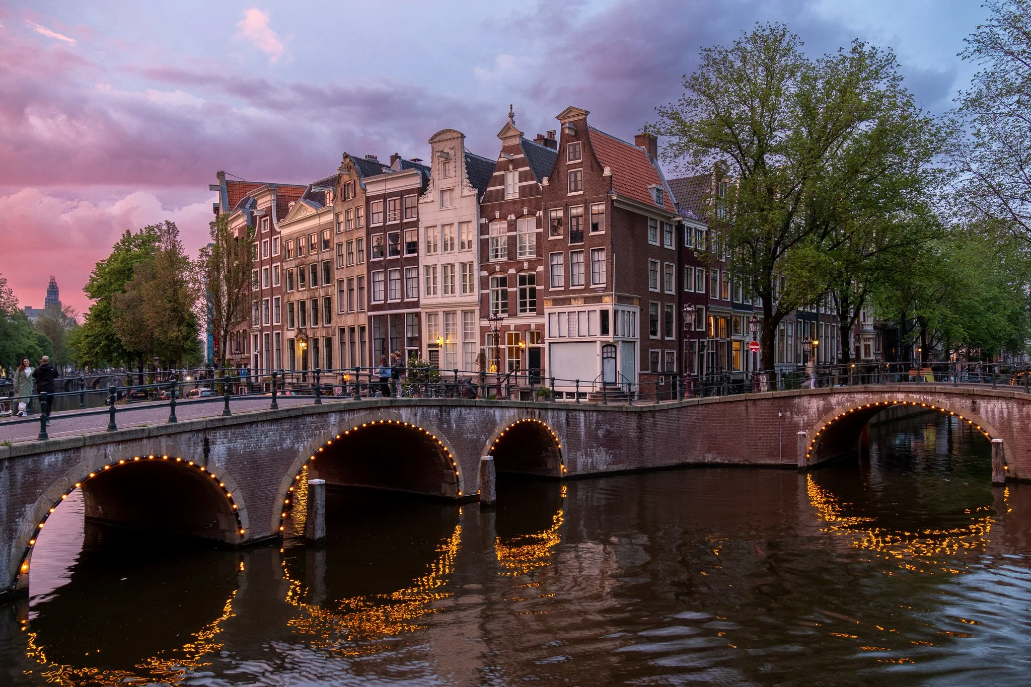 A canal in Amsterdam with traditional narrow brick houses and arched bridges illuminated by string lights at dusk.