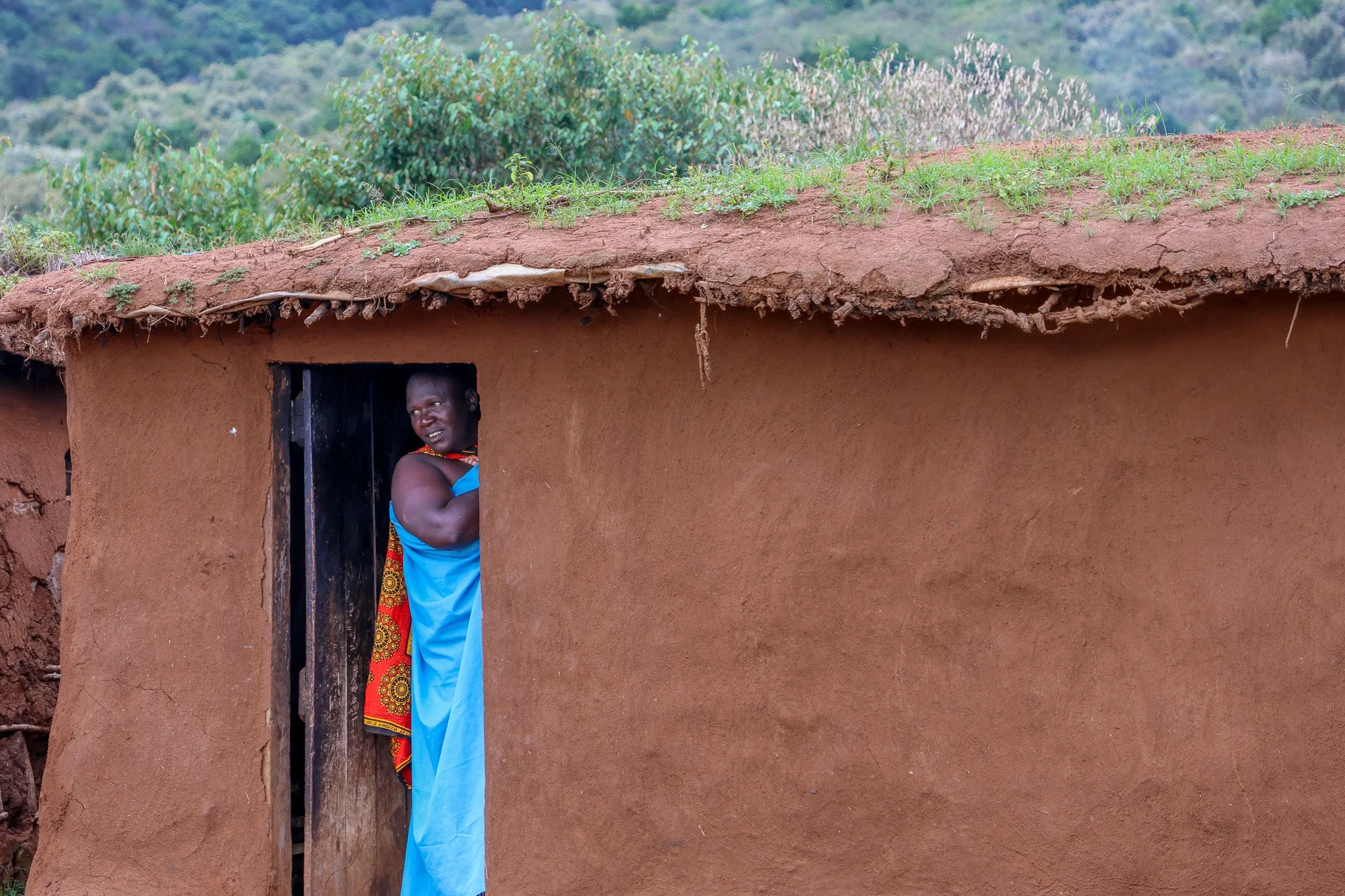 A woman in traditional attire standing in the doorway of a mud house with a green landscape in the background.