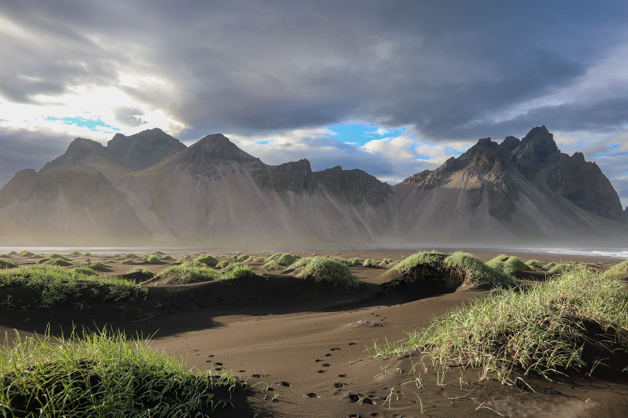 A scenic landscape of a mountainous region with dark clouds above, green grass-covered sand dunes in the foreground, and jagged mountains in the background.
