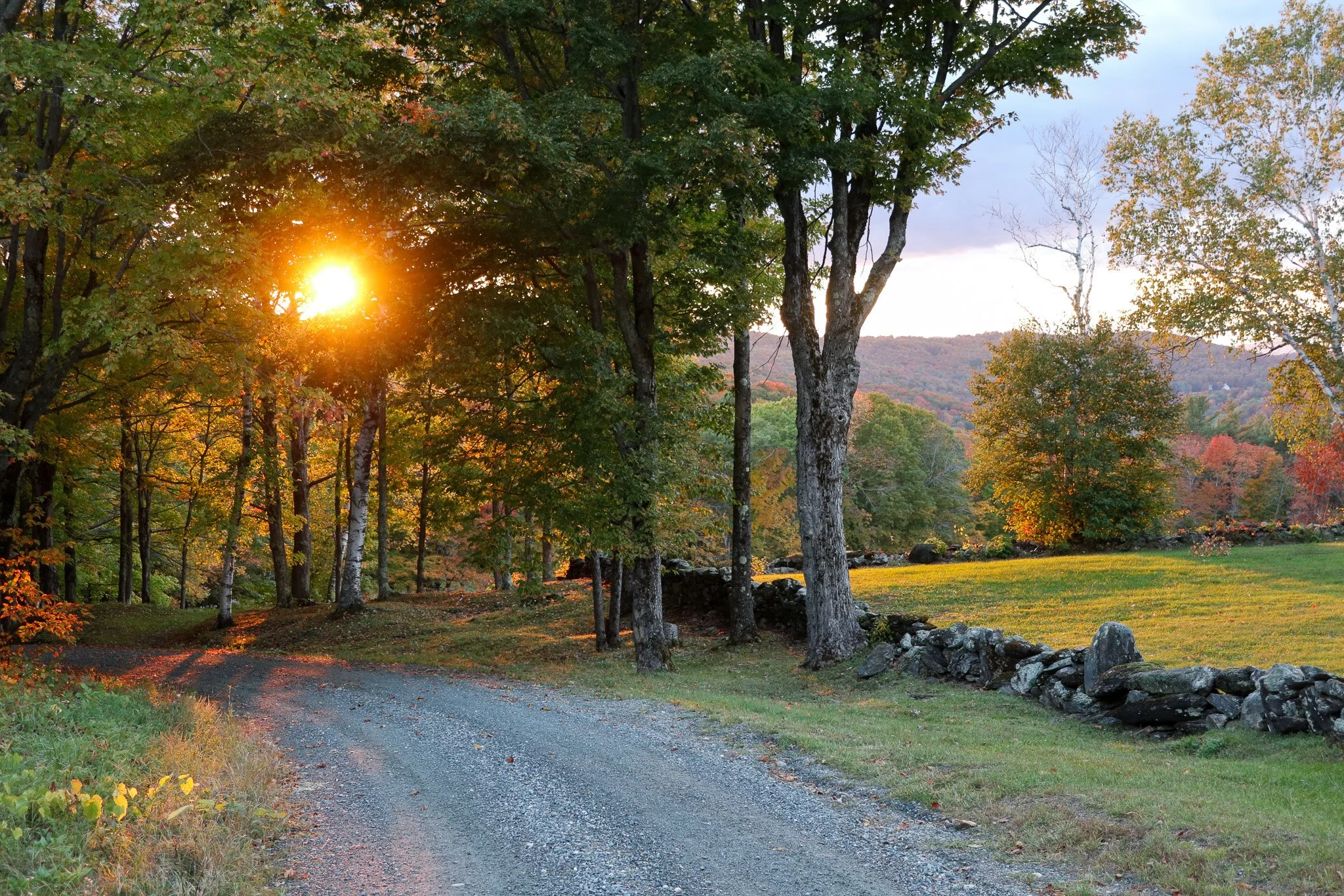 Sunset over a winding gravel road surrounded by trees with fall foliage and a stone wall on the right.