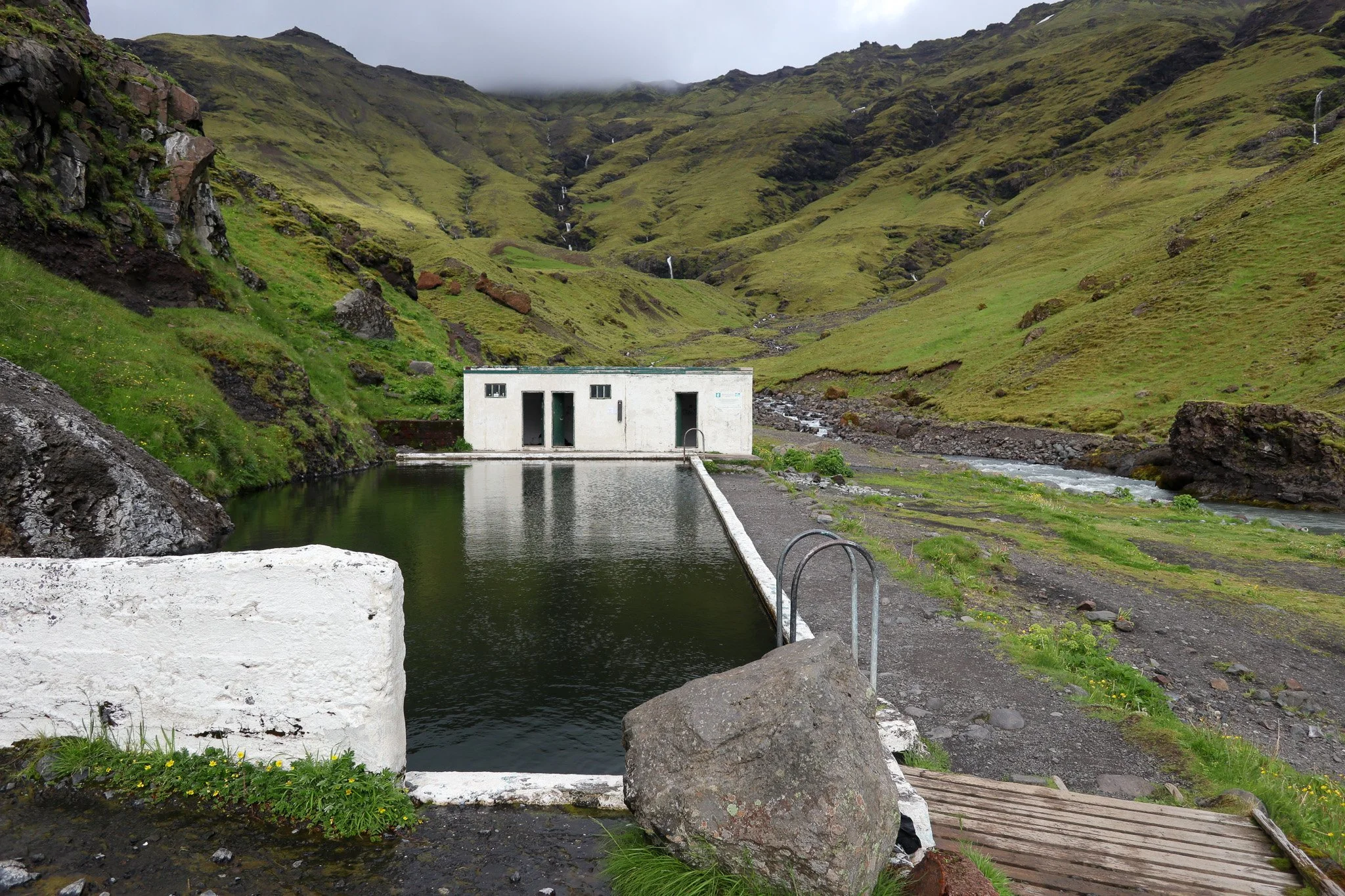 Small white building situated next to a rectangular pool of water, with green, moss-covered hills and streams in the background.