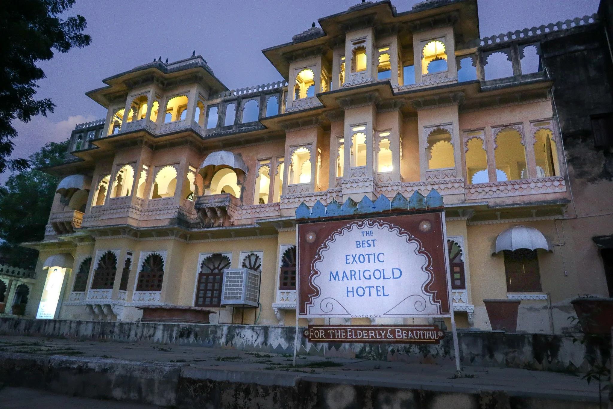 An old, multi-story hotel with ornate architecture and balcony railings, illuminated from the inside, with a sign in front reading "The Best Exotic Marigold Hotel," dedicated ‘For the Elderly & Beautiful,’ during dusk.