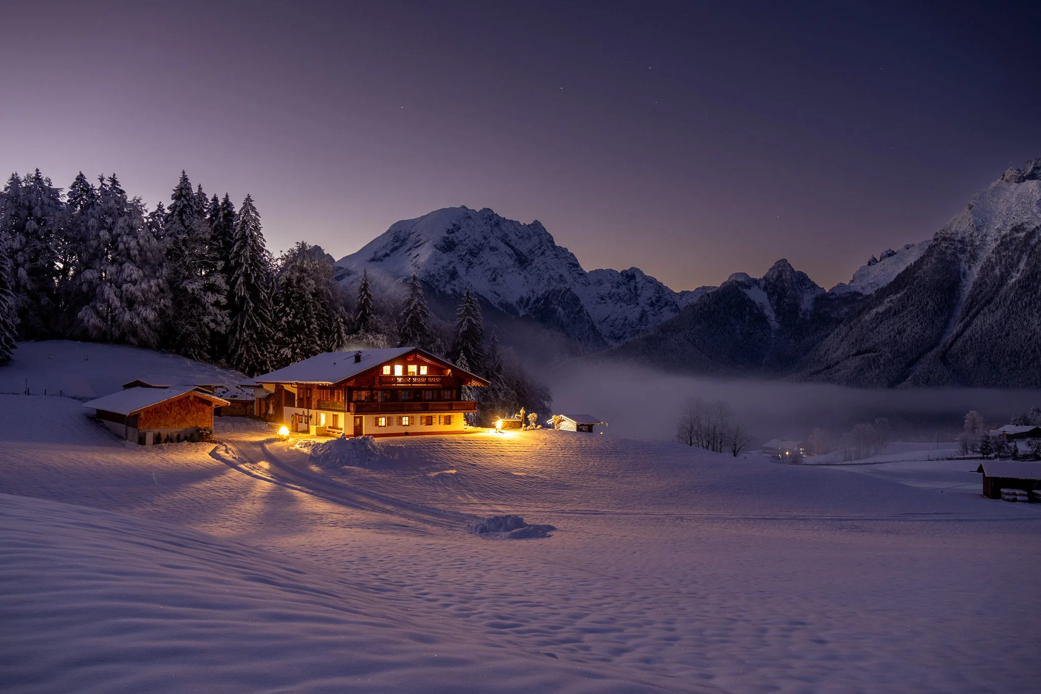 A house illuminated with warm lights in a snowy mountain landscape during twilight, with tall trees, distant mountains, and mist over a frozen lake.