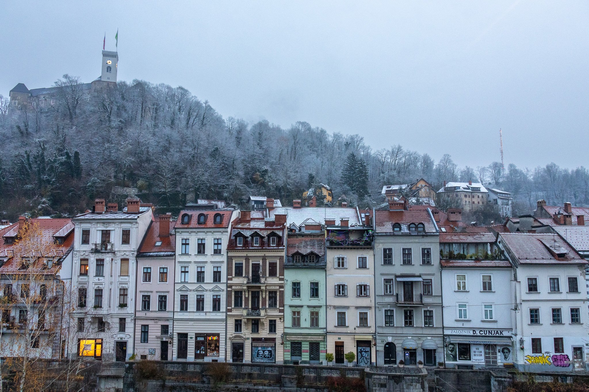 A cityscape with multi-story buildings featuring various architectural styles, balconies, and signs, set against a hillside with snowy trees and a castle-like structure with flags on top, in a cold, winter setting.