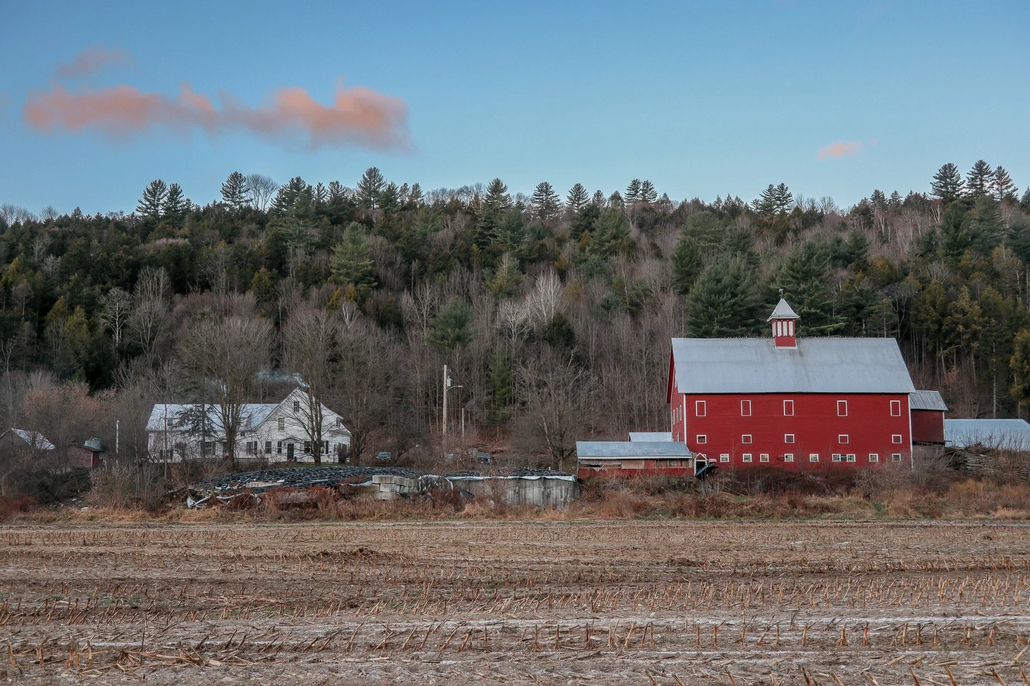 A rural farm scene with a red barn, a white farmhouse, trees, and a cloudy blue sky.
