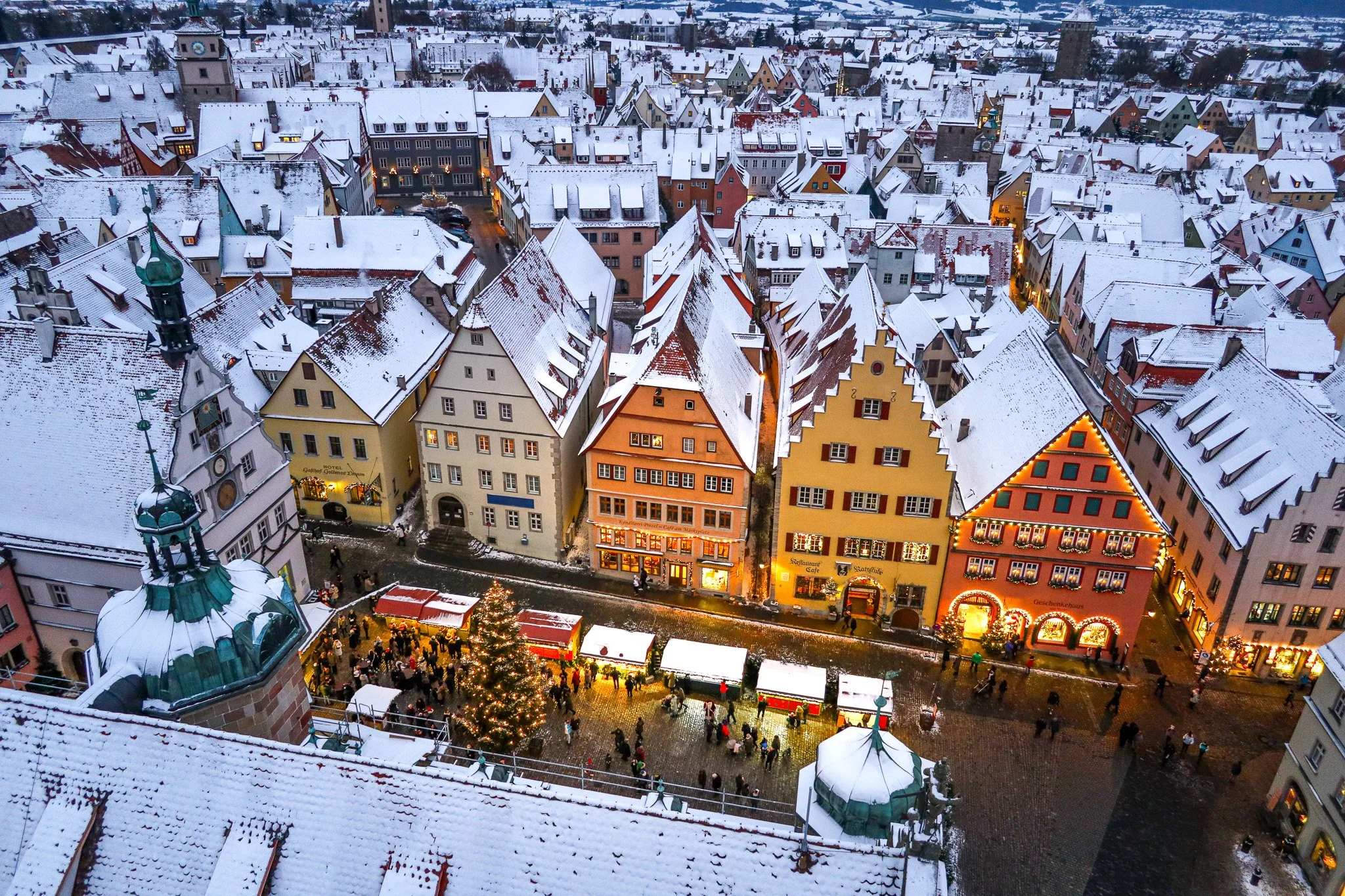 A snowy aerial view of a European town square with colorful buildings decorated for Christmas, a lit Christmas tree, and market stalls with people walking around in winter.