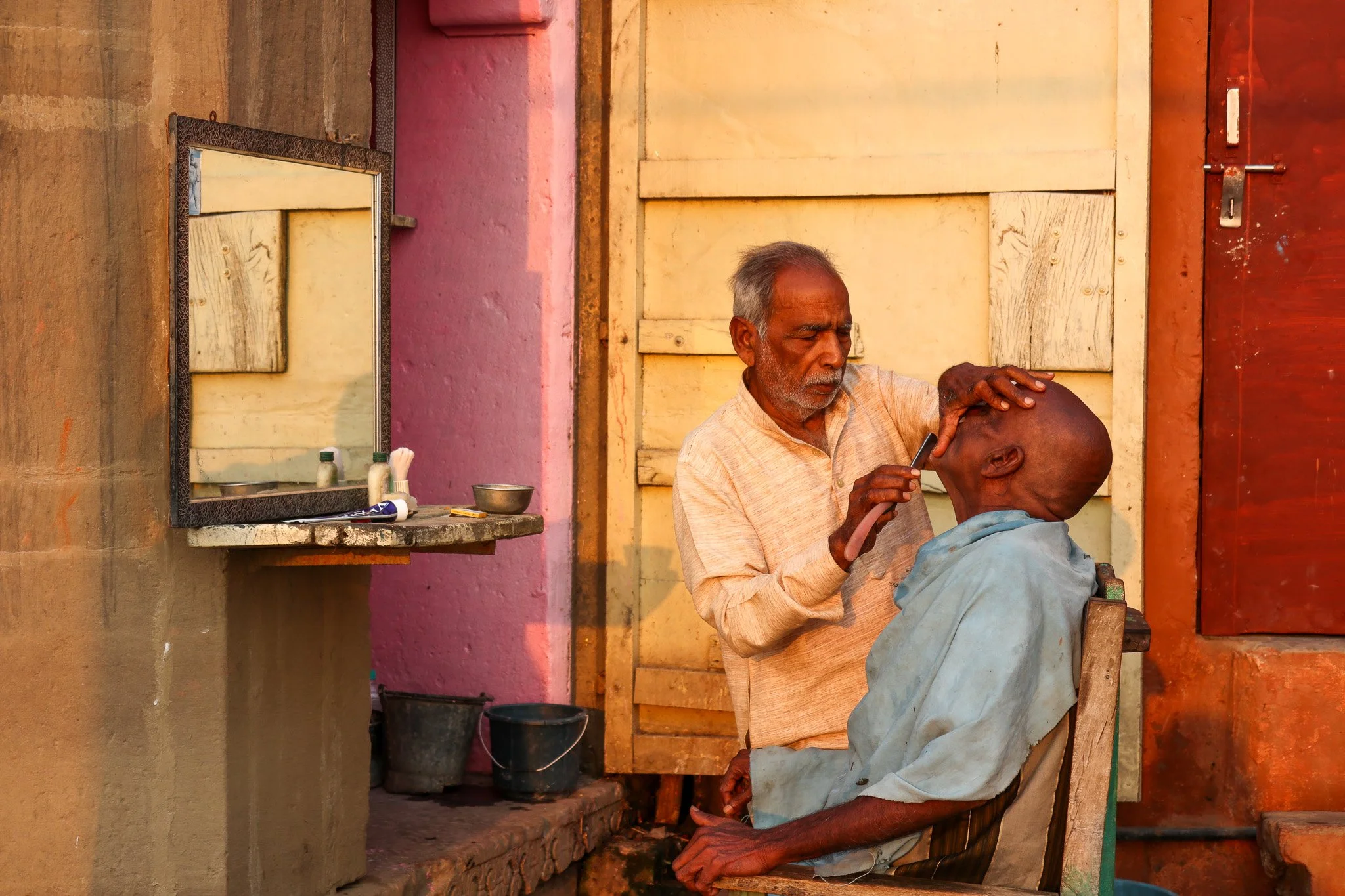 An elderly man getting a shave at an outdoor barber shop with a man shaving his head.