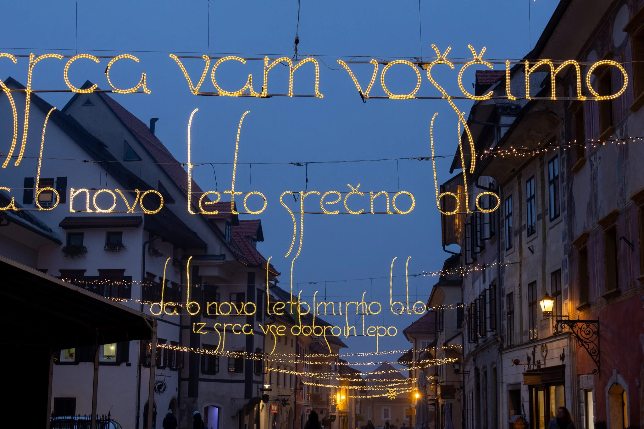 Street decorated with glowing holiday lights in the evening, with a greeting in a foreign language suspended above.