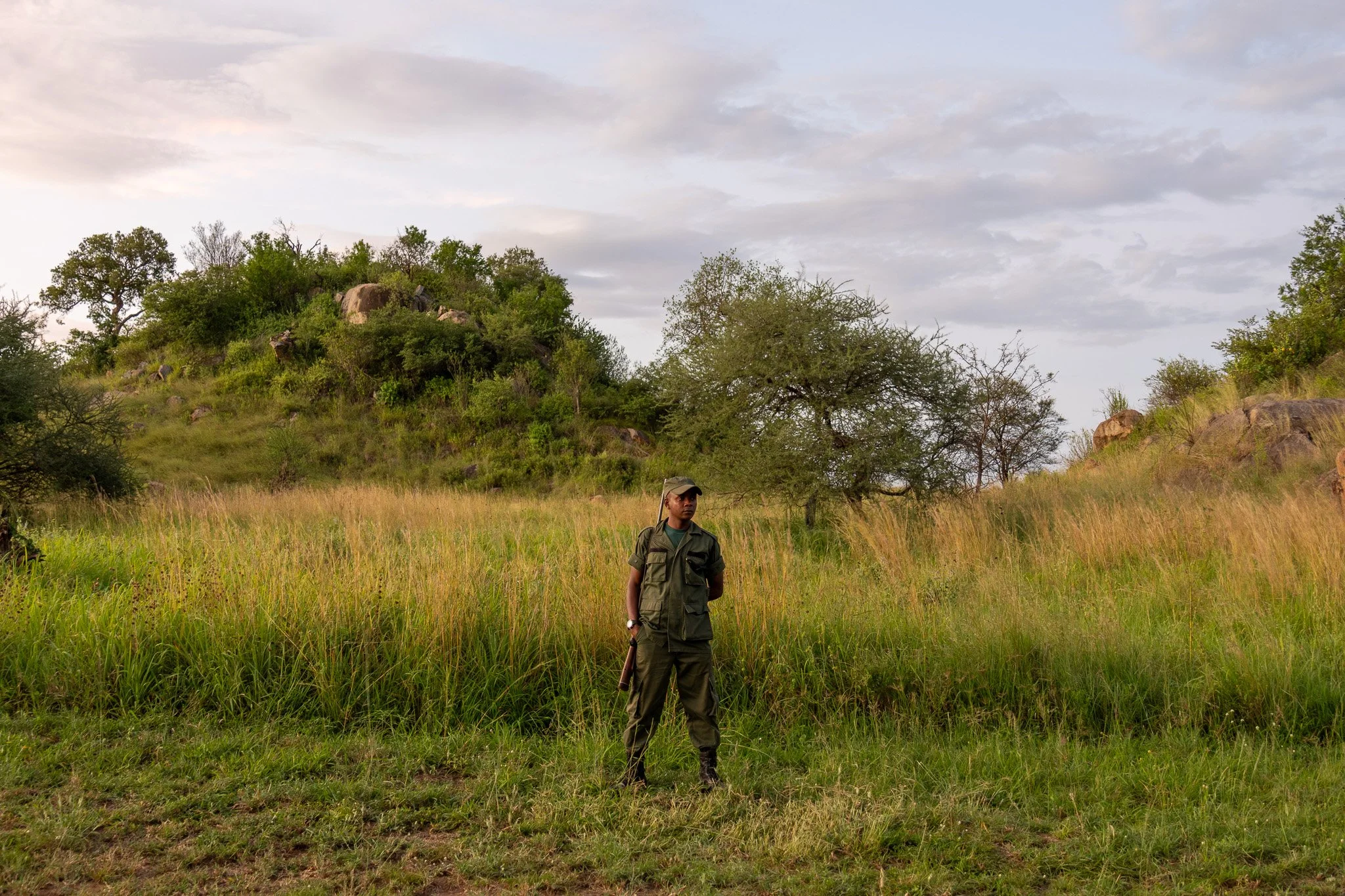 A person in a green outfit standing in a grassy field with trees and hills in the background.