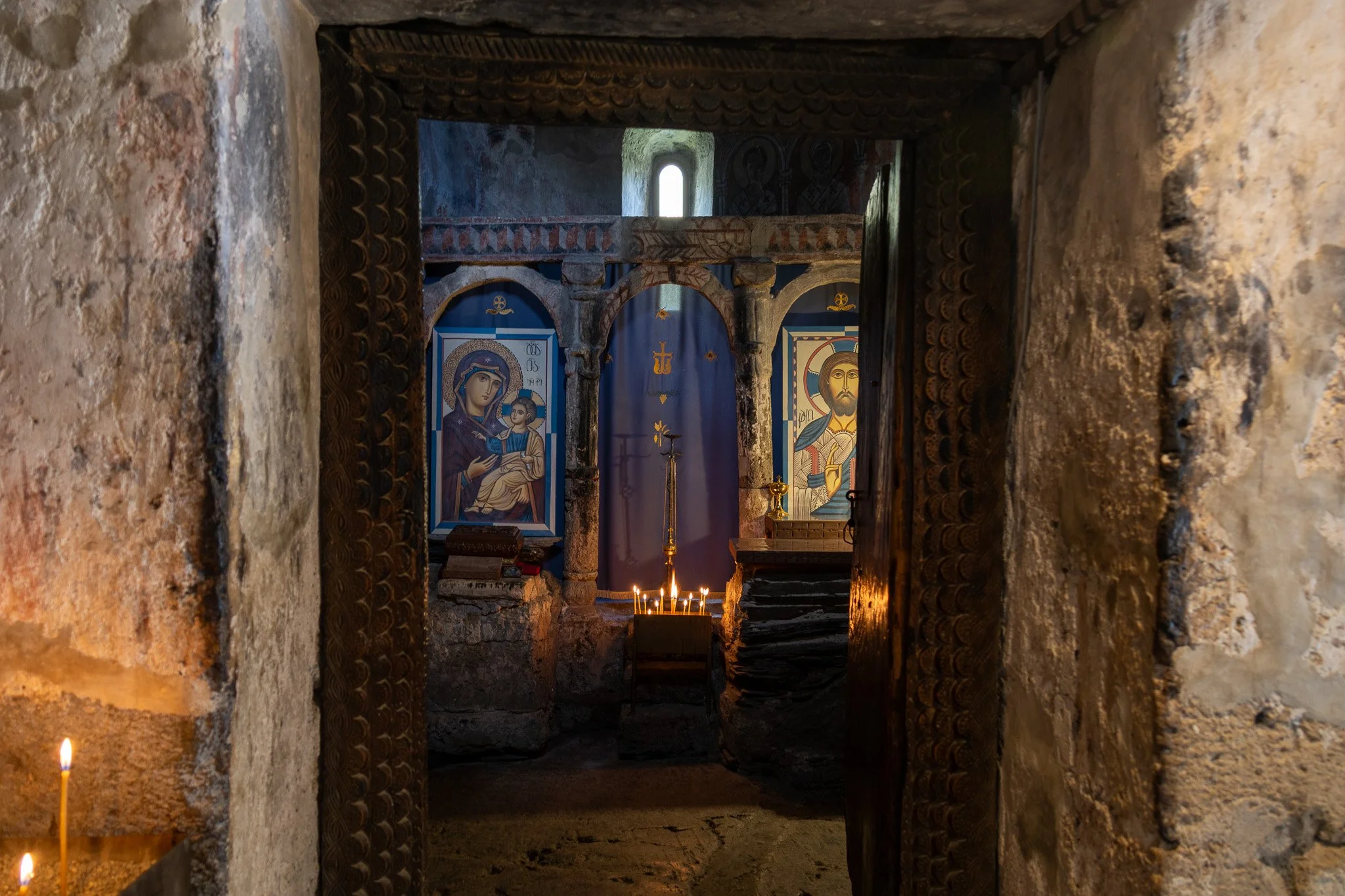 View through a doorway into a small, dimly-lit religious chapel featuring icons of Mary and Jesus, lit candles, and stone walls.