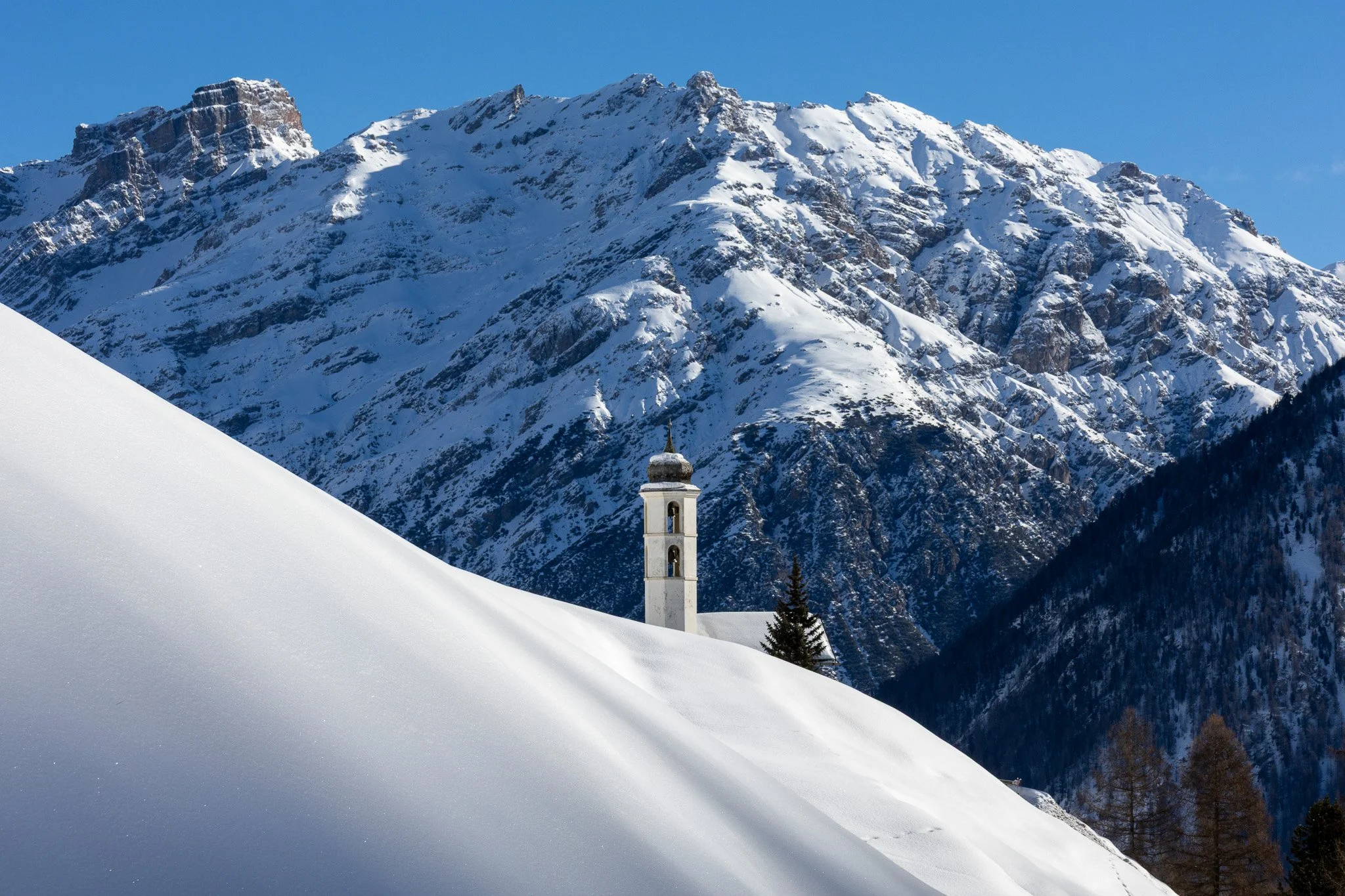 Snow-covered mountain landscape with a church steeple and tall trees in the foreground