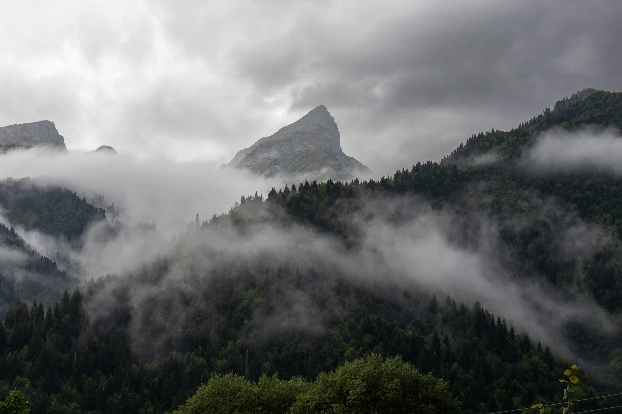 Cloudy mountain landscape with fog rolling over dense forested hills and a prominent peak in the background.