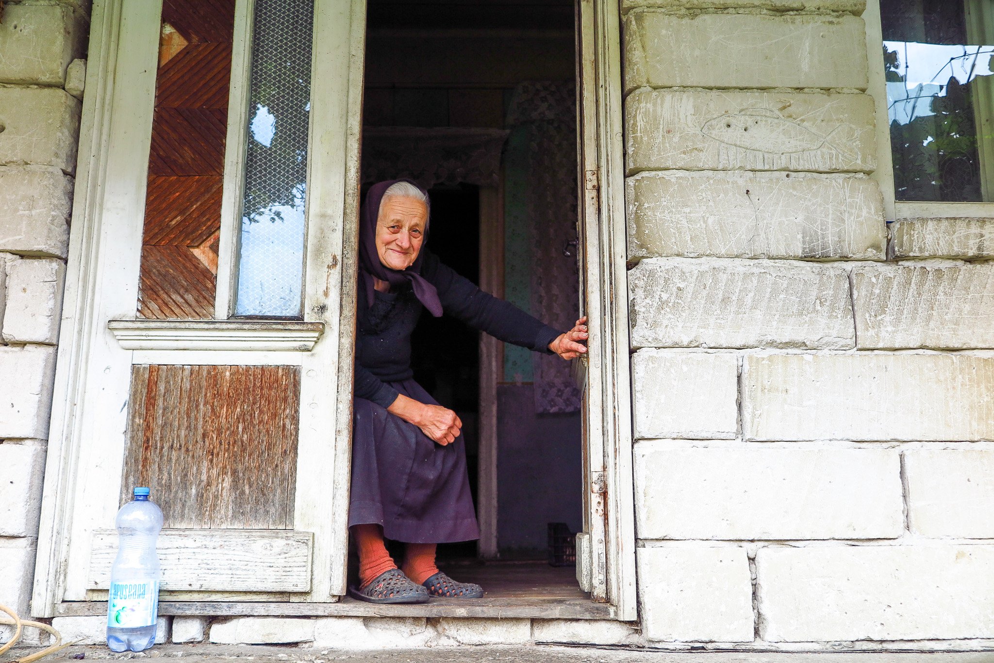 An elderly woman sitting at her doorway, smiling and holding the door frame, with a bottle of water on the step outside.