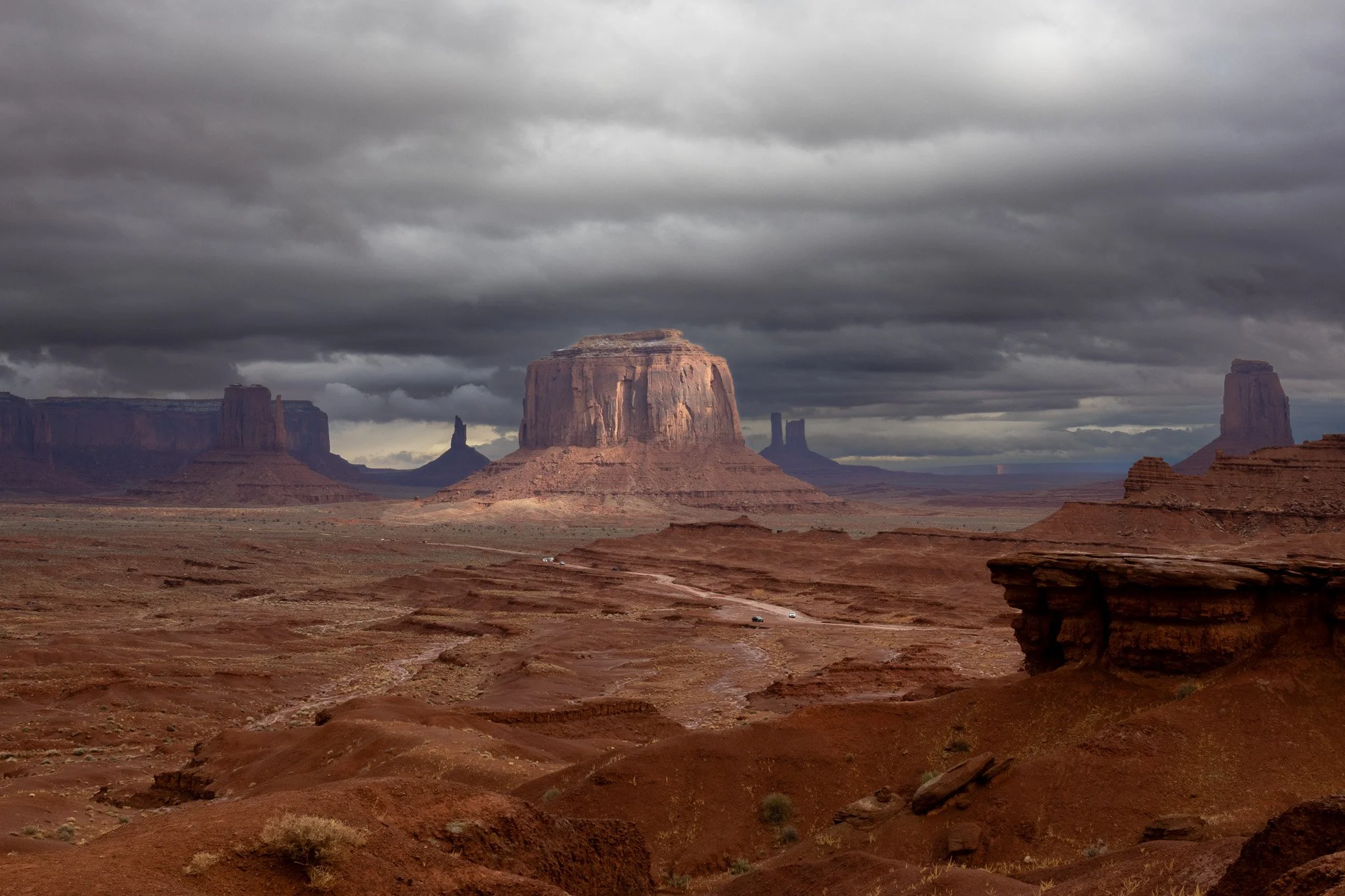 A wide view of Monument Valley showing large sandstone buttes under a cloudy sky, with a dirt road and vehicles in the foreground.