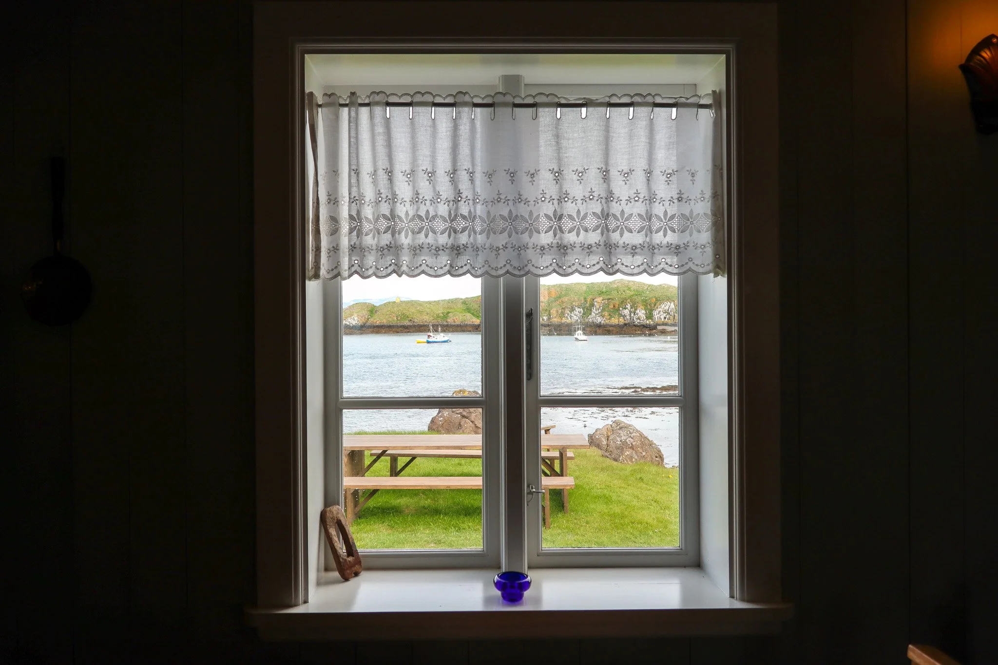 View through a window showing a grassy area, rocks, water, and boats in the distance, with a wooden picnic table outside.