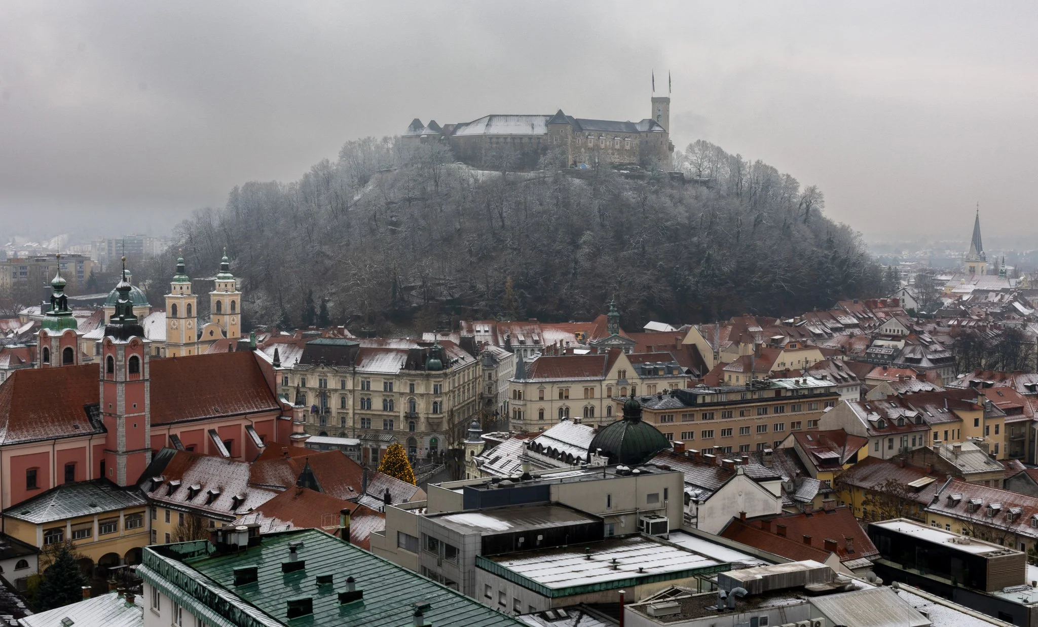 A scenic view of a European town with rooftops covered in snow, churches with tall steeples, and a castle on a hill surrounded by snow-covered trees on a cloudy winter day.