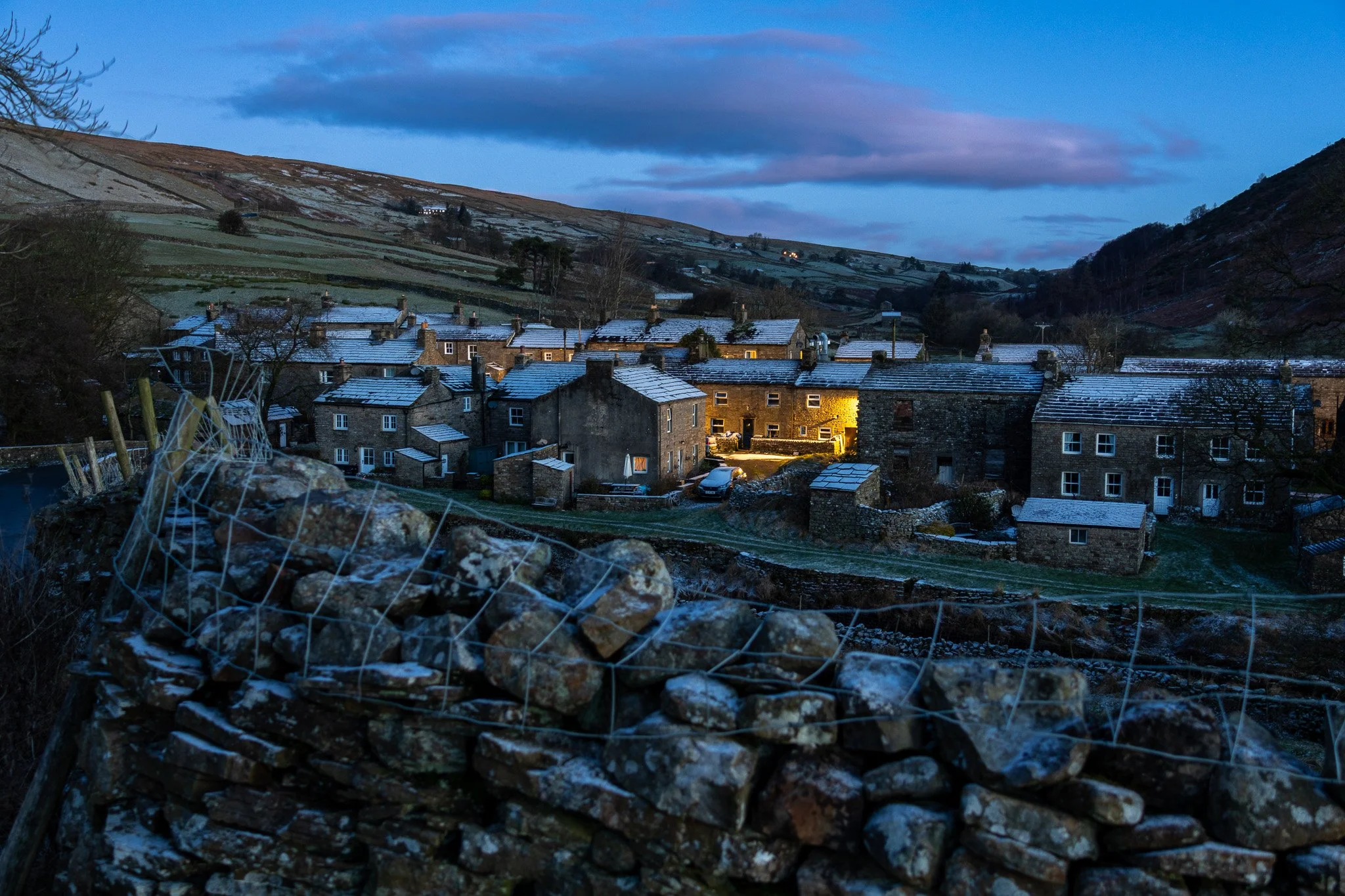 A small village at dusk, with snow on rooftops and hills in the background, and a stone wall with a wire fence in the foreground.