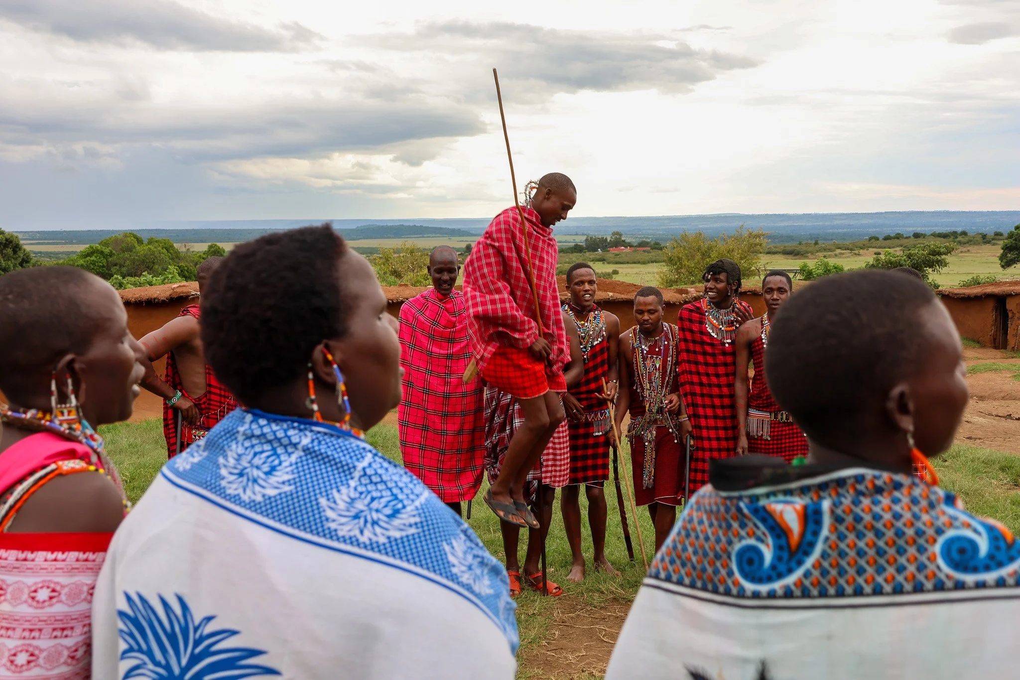 A group of Maasai people wearing traditional red and blue shuka clothing, with beaded jewelry, gathered outdoors in a rural setting with trees and fields in the background. One Maasai man is jumping in the air while others watch and smile.