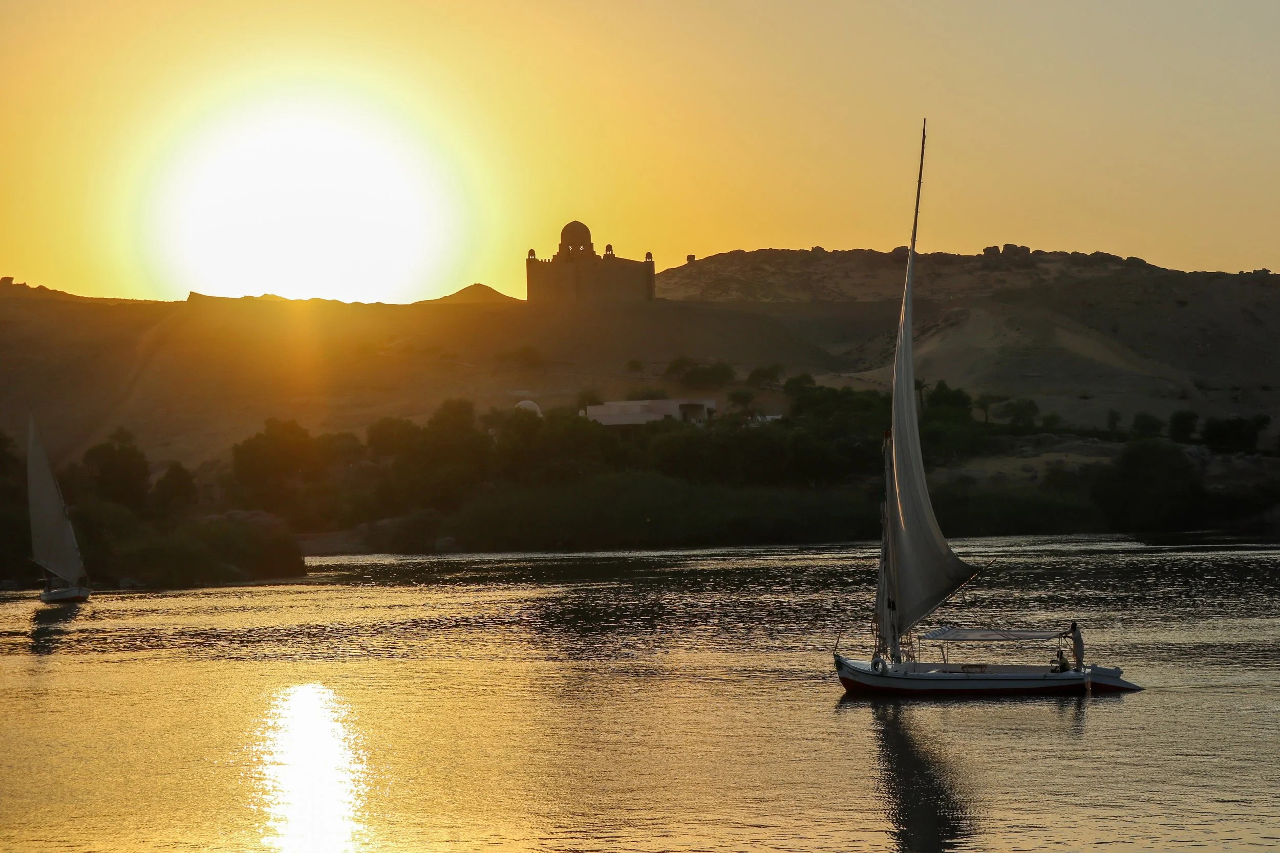 A sailboat on calm water during sunset with a hill and a building with domes in the background.