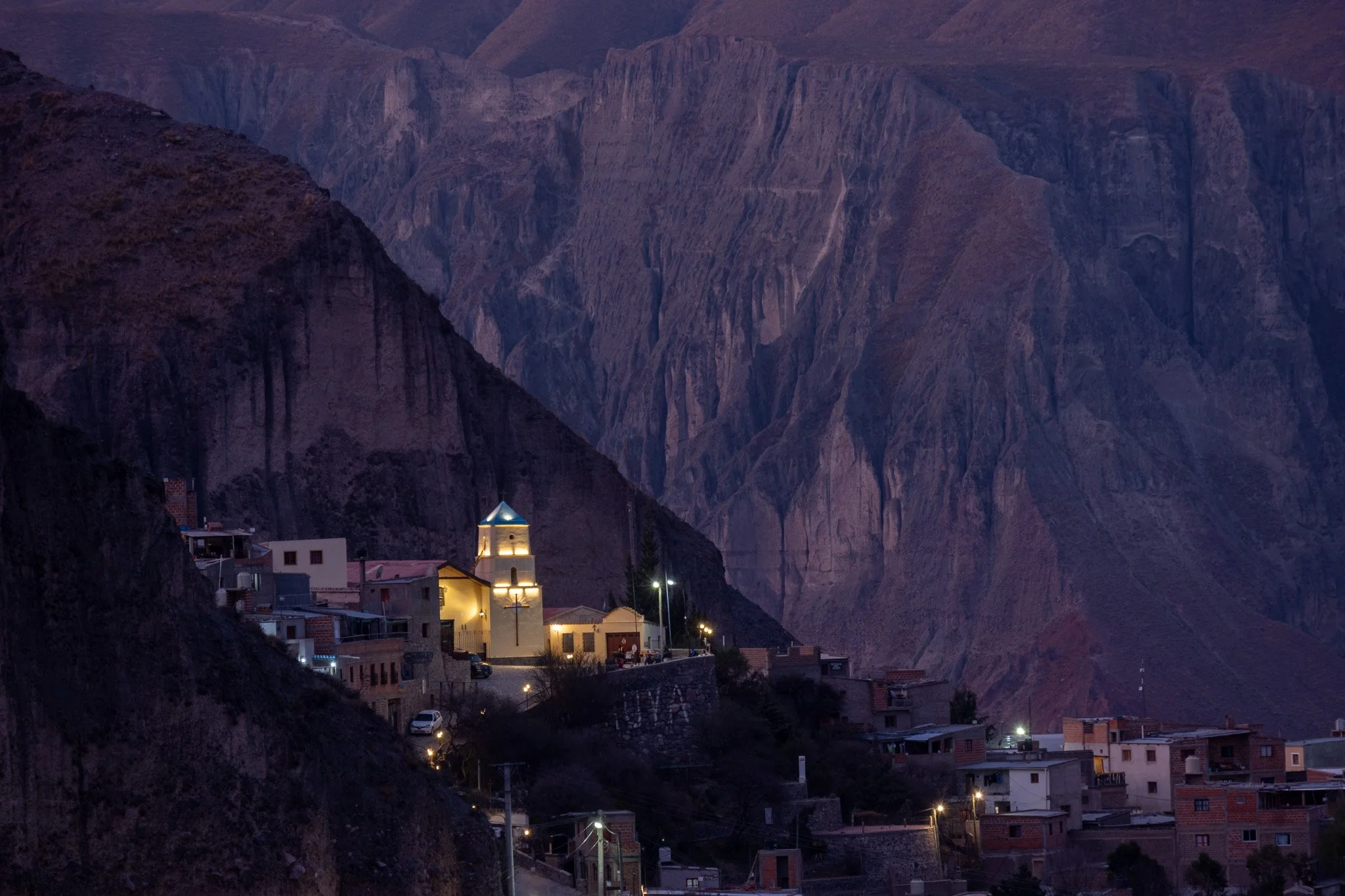 Small town at the base of rugged mountain cliffs during dusk, with illuminated buildings including a church with a blue roof.