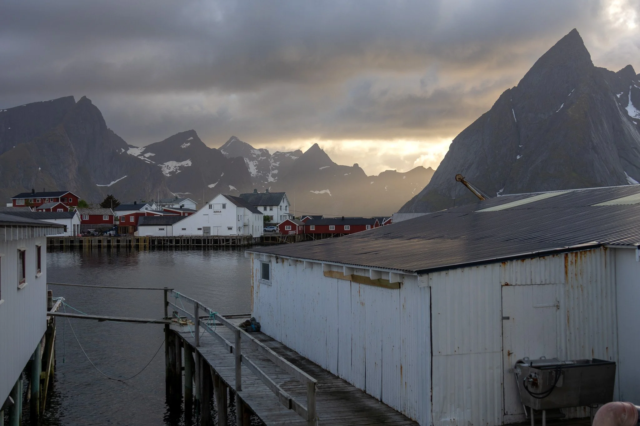 A coastal village with white and red houses along the water, surrounded by tall, rugged mountains under a partly cloudy sky during sunset or sunrise.