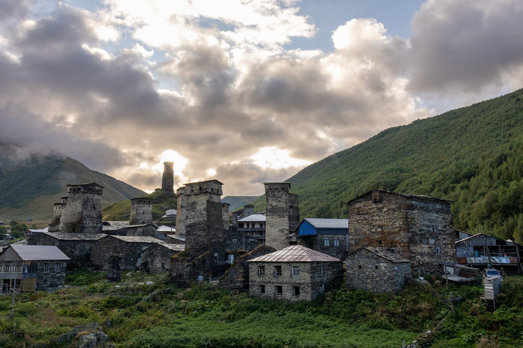 Stone towers and buildings in a mountain village with greenery, under cloudy sky at sunset.