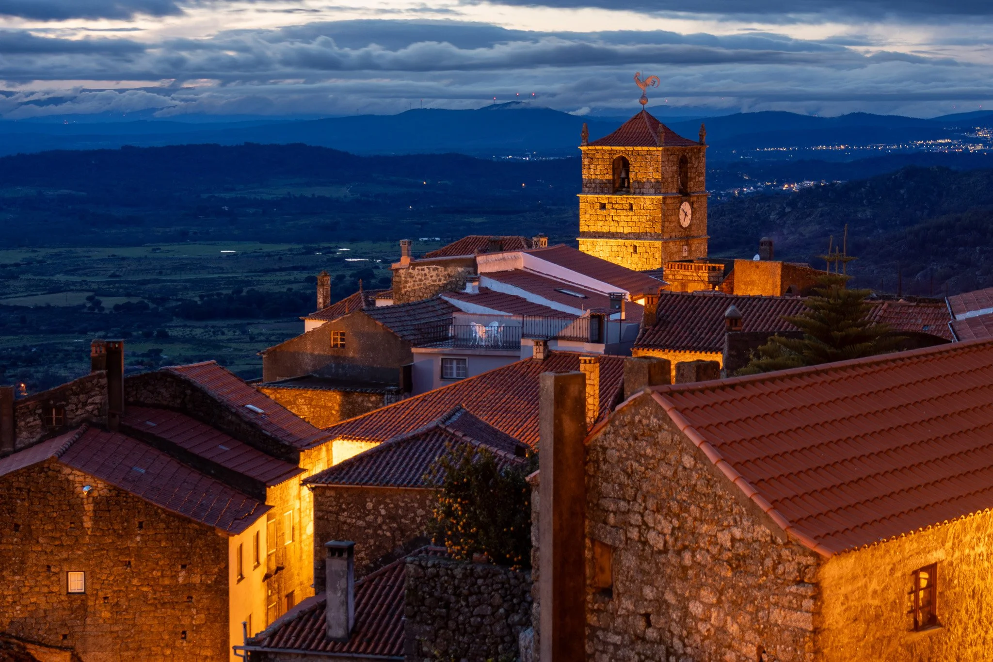 A scenic view of a small European town with stone buildings and red-tiled roofs illuminated by warm sunset light, featuring a prominent clock tower with a weather vane on top, set against rolling hills and a cloudy sky at dusk.