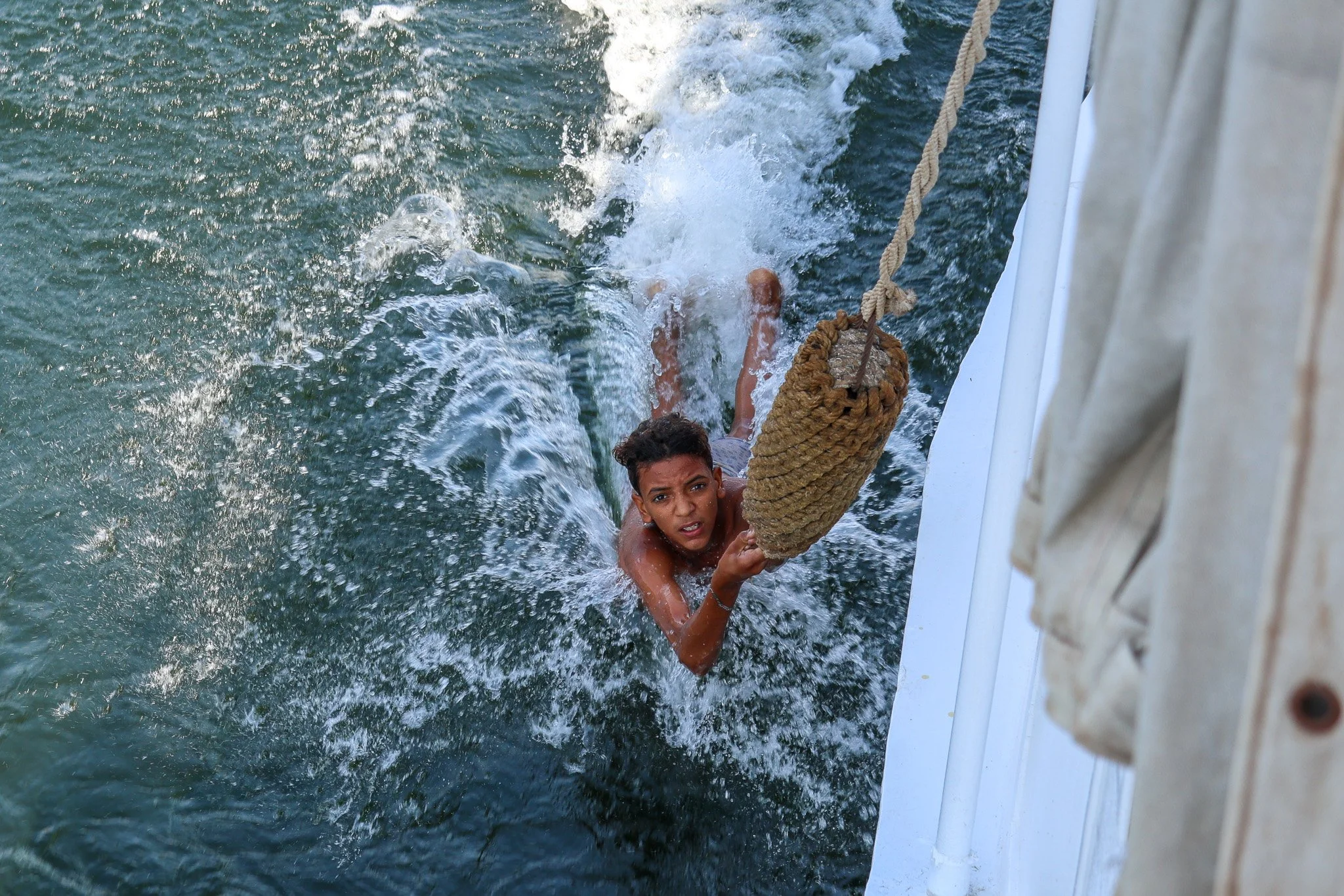 A boy is holding onto a rope while hanging off the side of a boat, with water splashing around him as he is in the water.