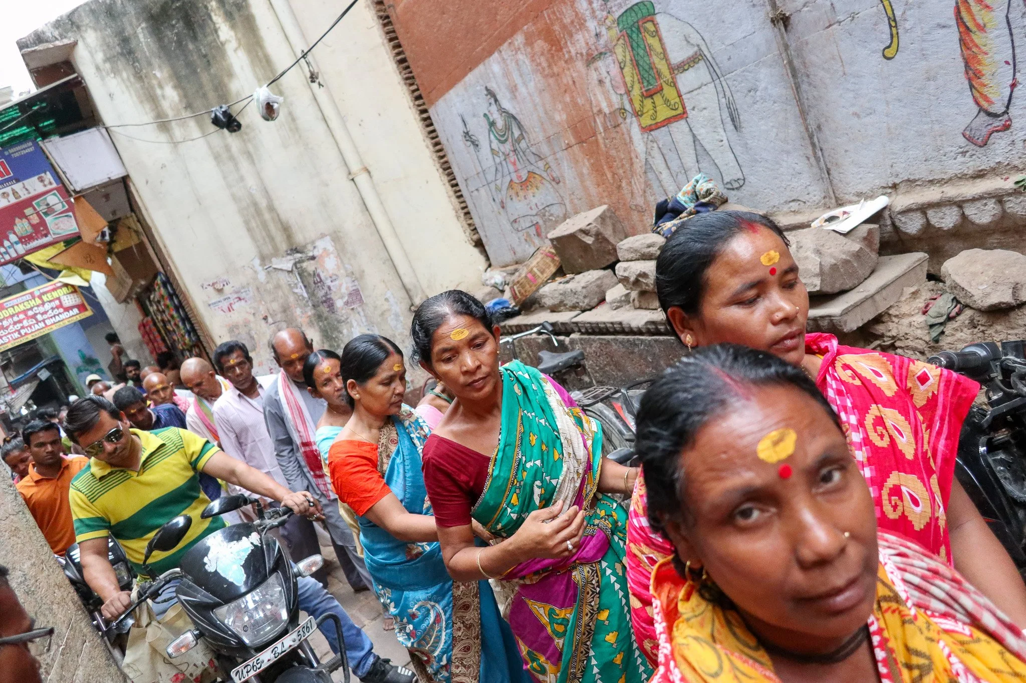 Line of people, mainly women in colorful traditional clothing and some men, standing near motorcycles and on a narrow street with faded walls and murals in the background.