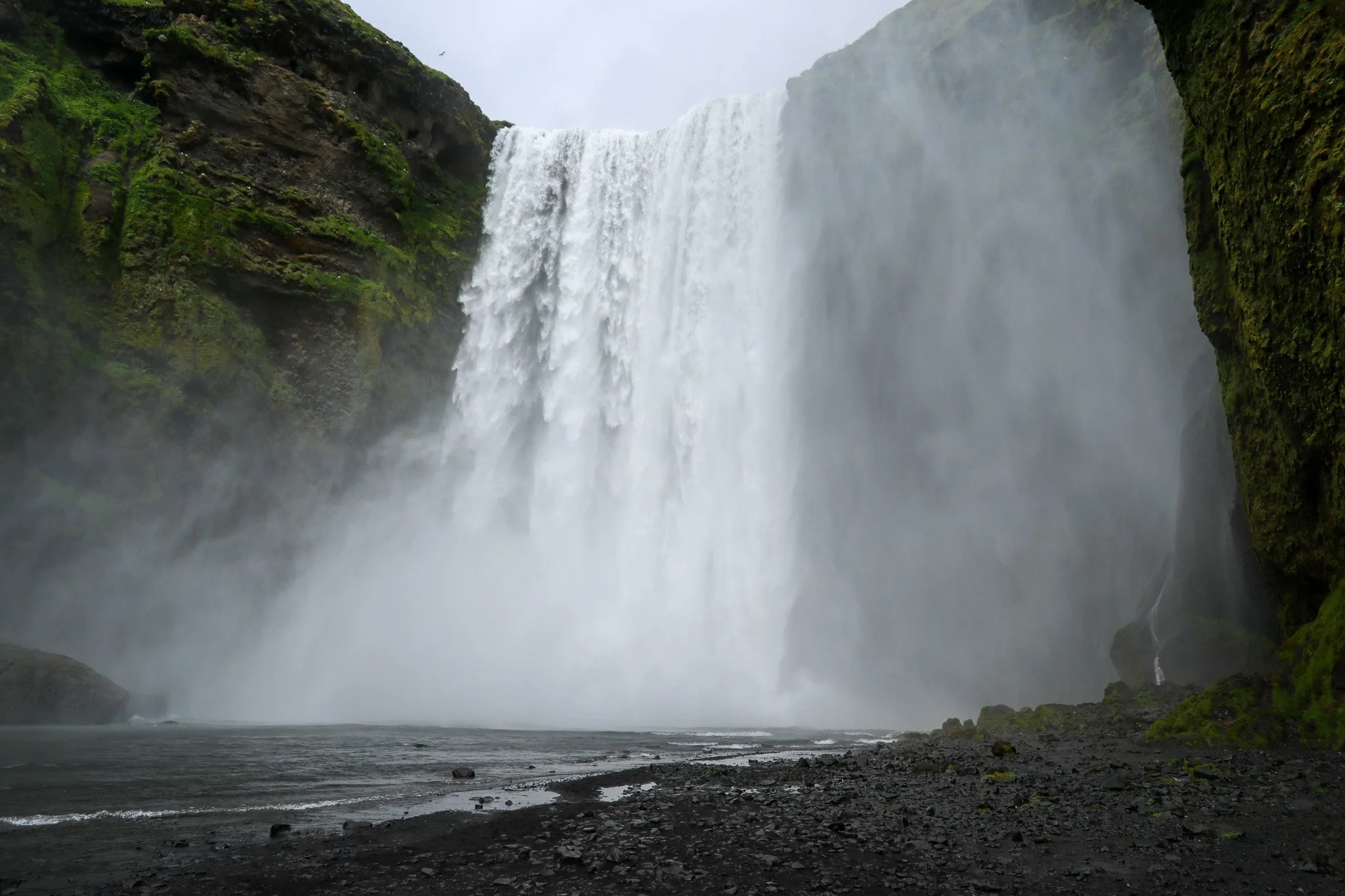 A large waterfall cascading down Moss-covered cliffs into a rocky river below, with mist rising at the base.