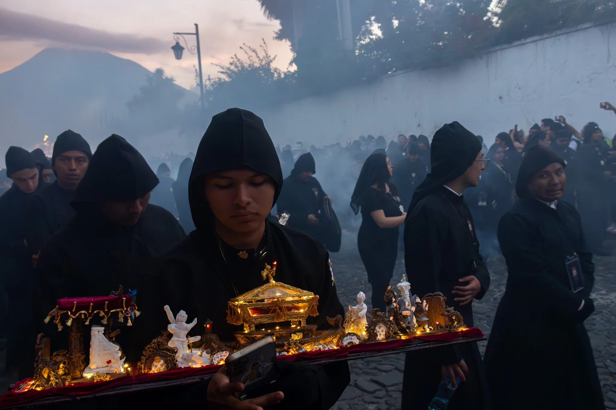 People dressed in black clothing and hooded garments participating in a solemn outdoor procession or ceremony, carrying a tray with religious or cultural figurines and decorative objects, with a misty background and a mountain visible in the distance