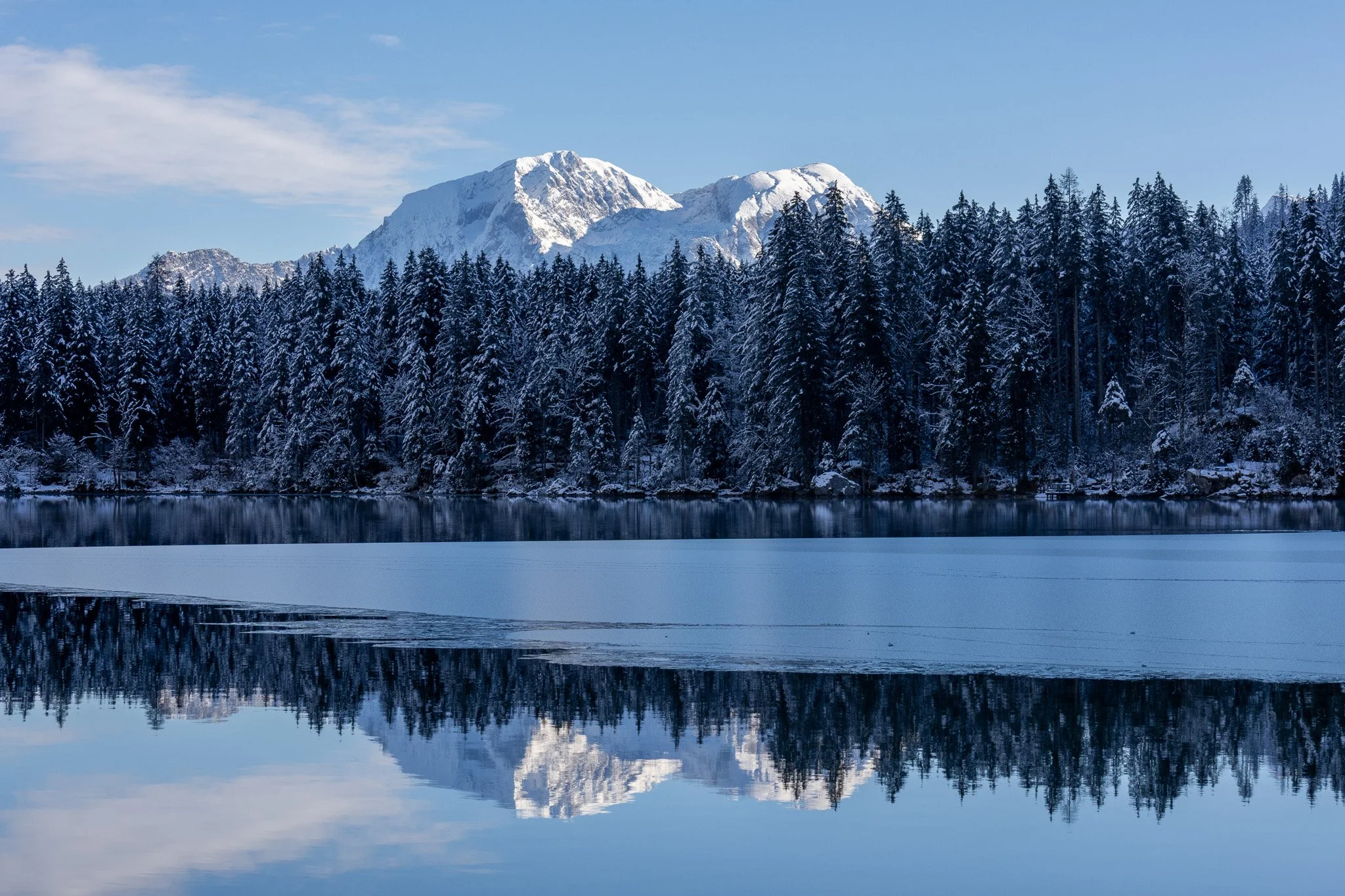 Snow-covered mountains and dense pine forest reflected on a partially frozen lake under a clear blue sky.